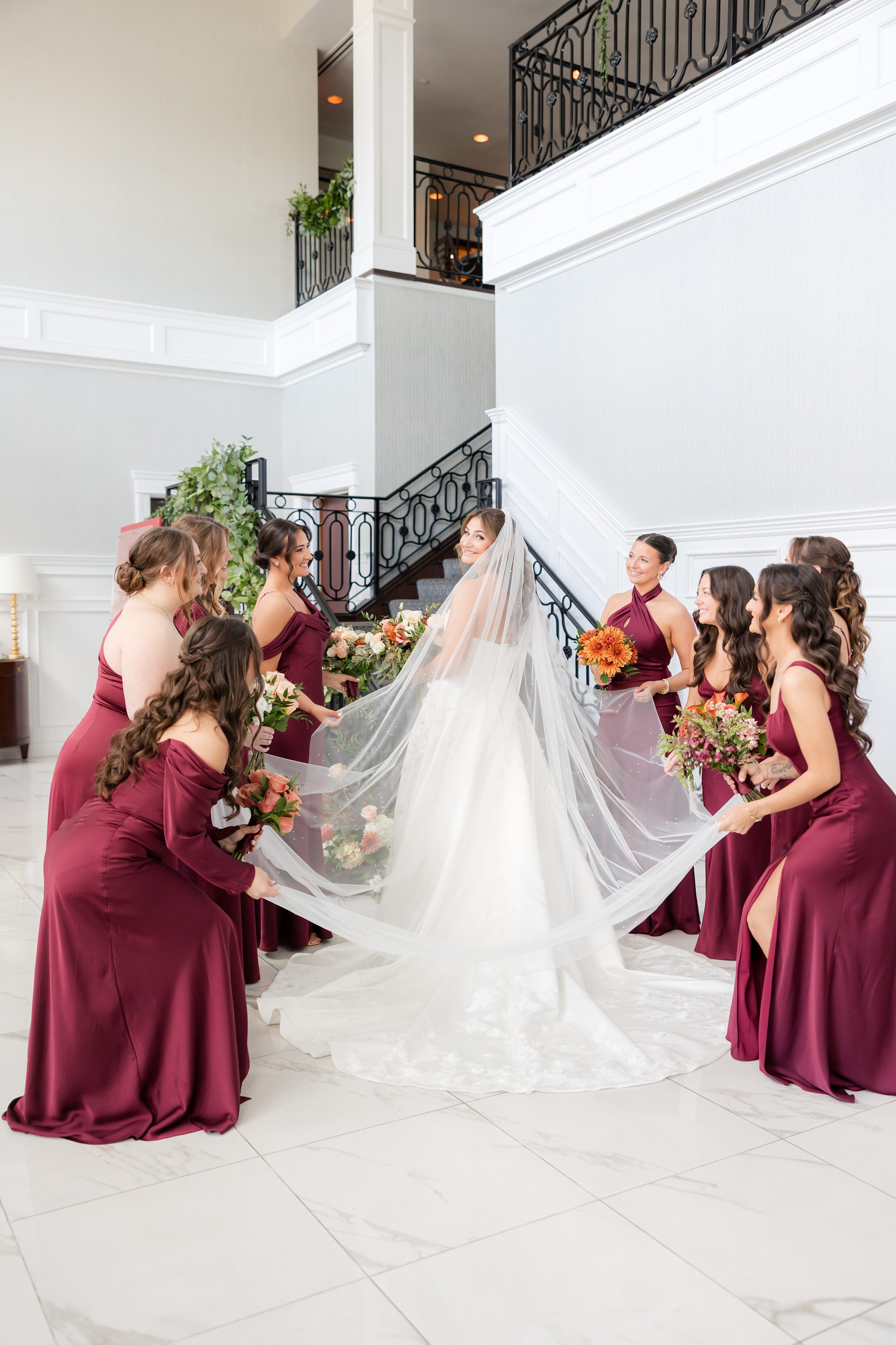 Bride surrounded by bridesmaids in matching burgundy dresses, holding her veil indoors.