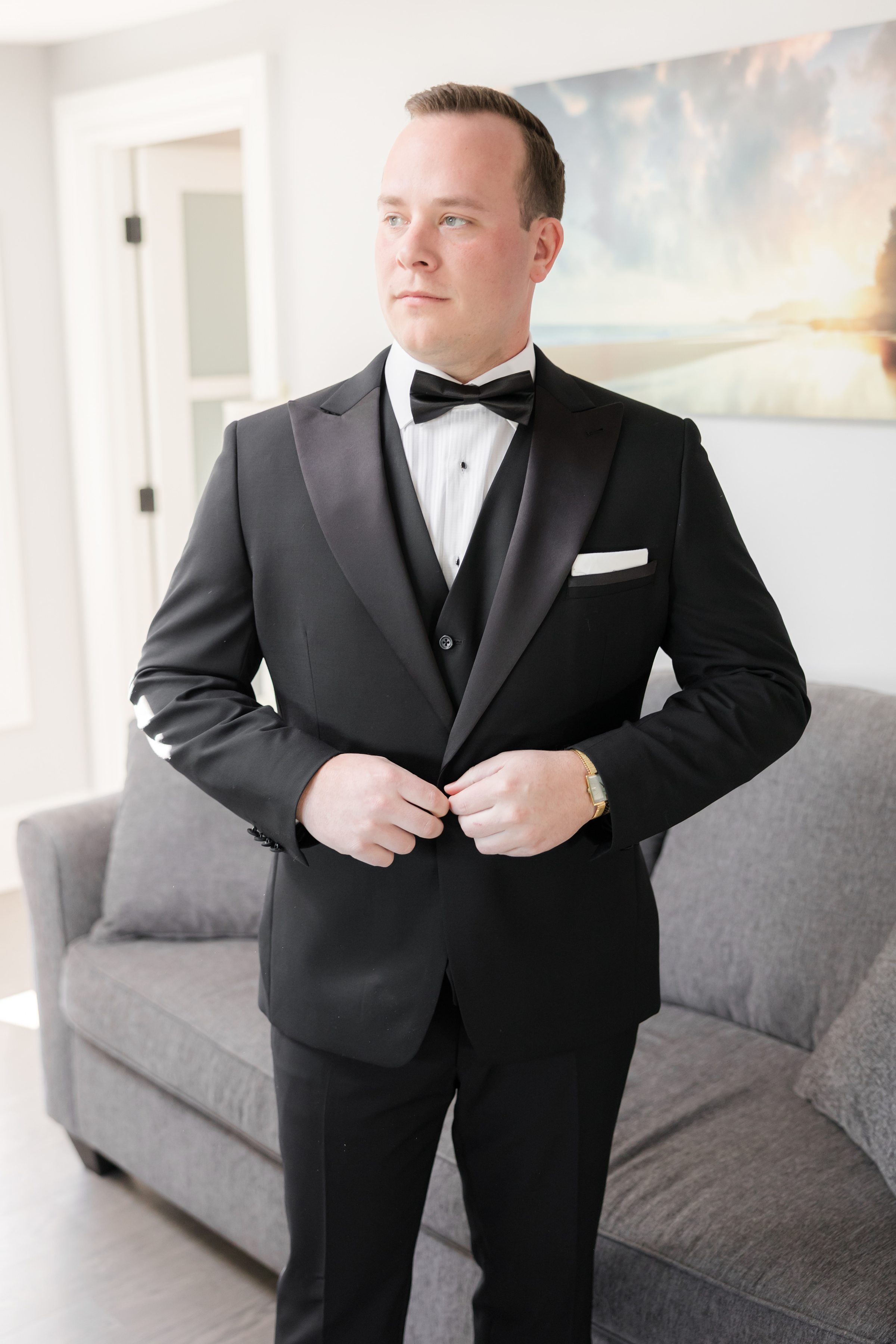 Groom in a classic black tuxedo fastening his jacket indoors, gazing off to the side in soft natural light.