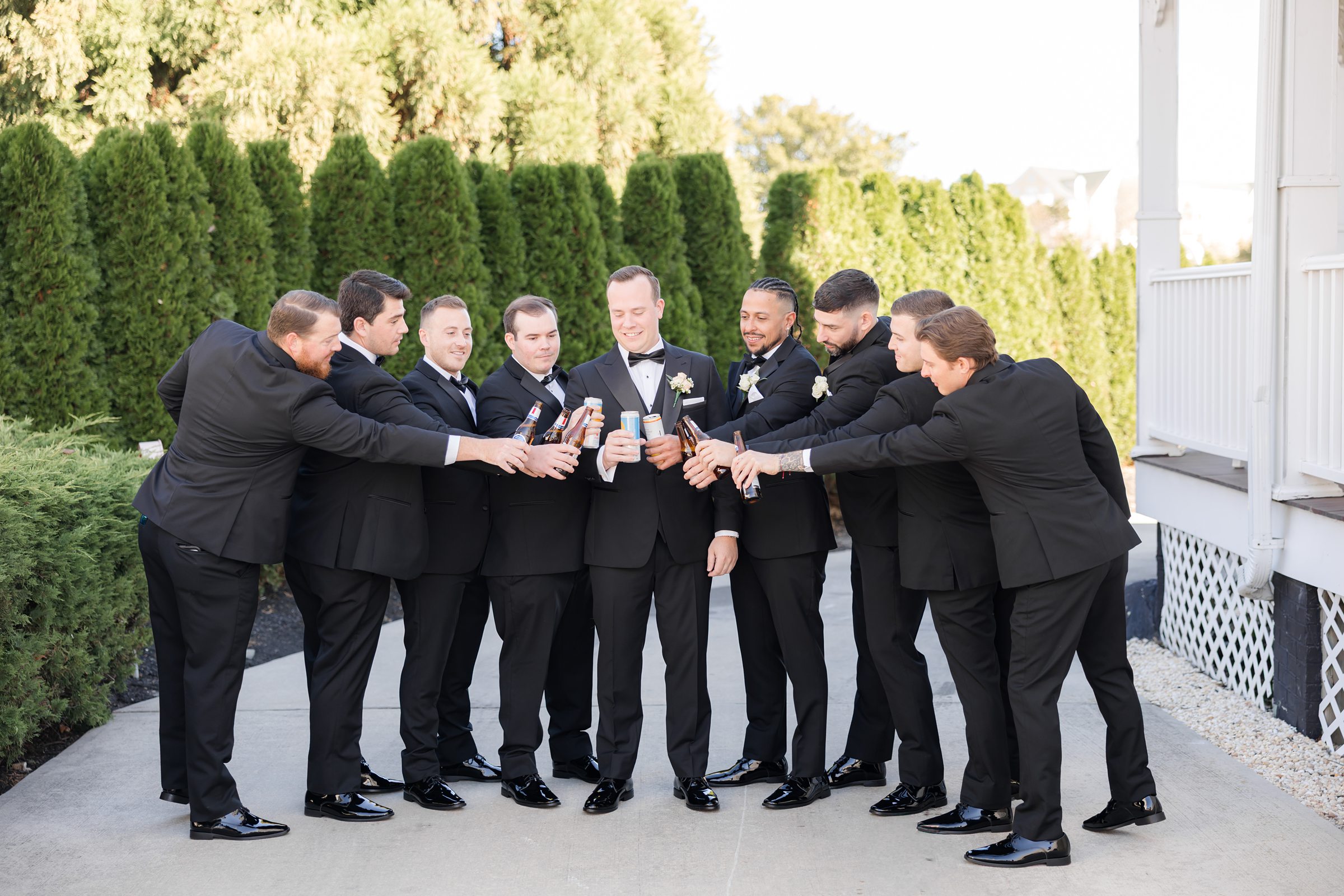 Groom and groomsmen in black tuxedos clinking drinks together outdoors