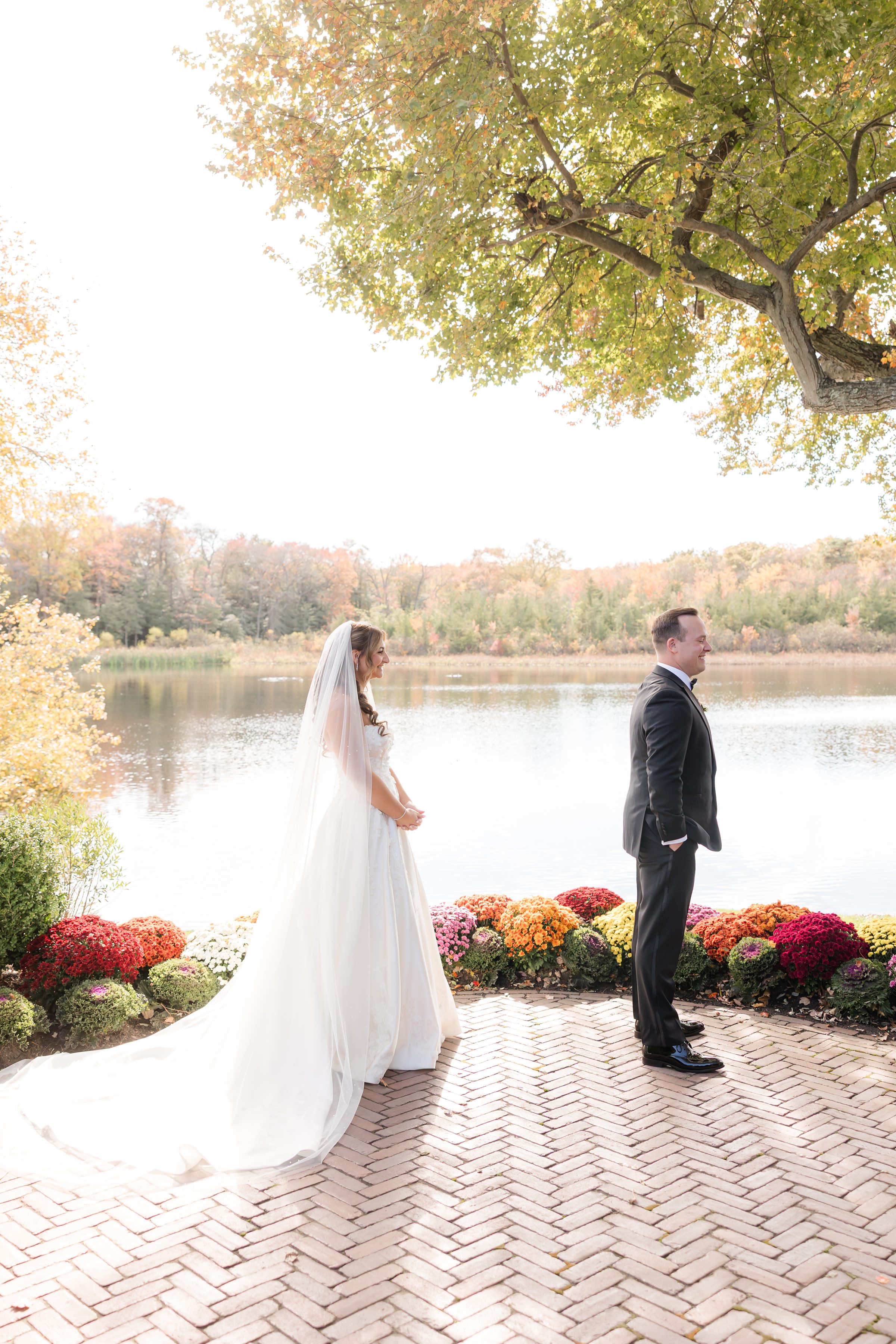 Bride and groom share a quiet first-look moment by a serene lakeside, surrounded by soft autumn colors and blooming flowers.