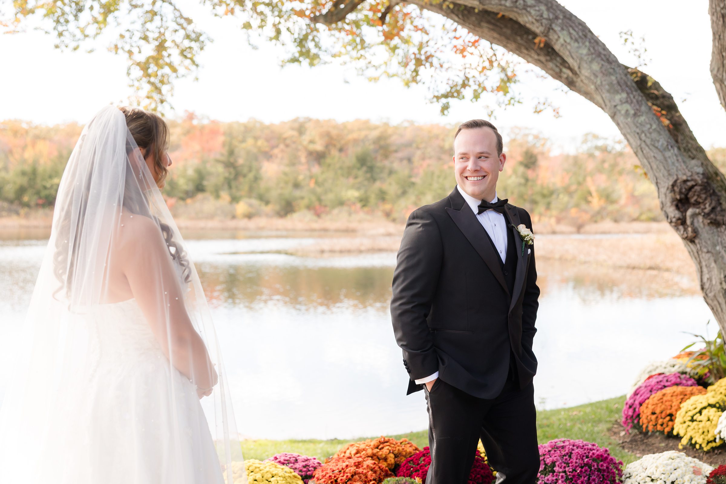 Groom smiling warmly as he turns toward the bride during their lakeside first look, surrounded by vibrant autumn flowers.