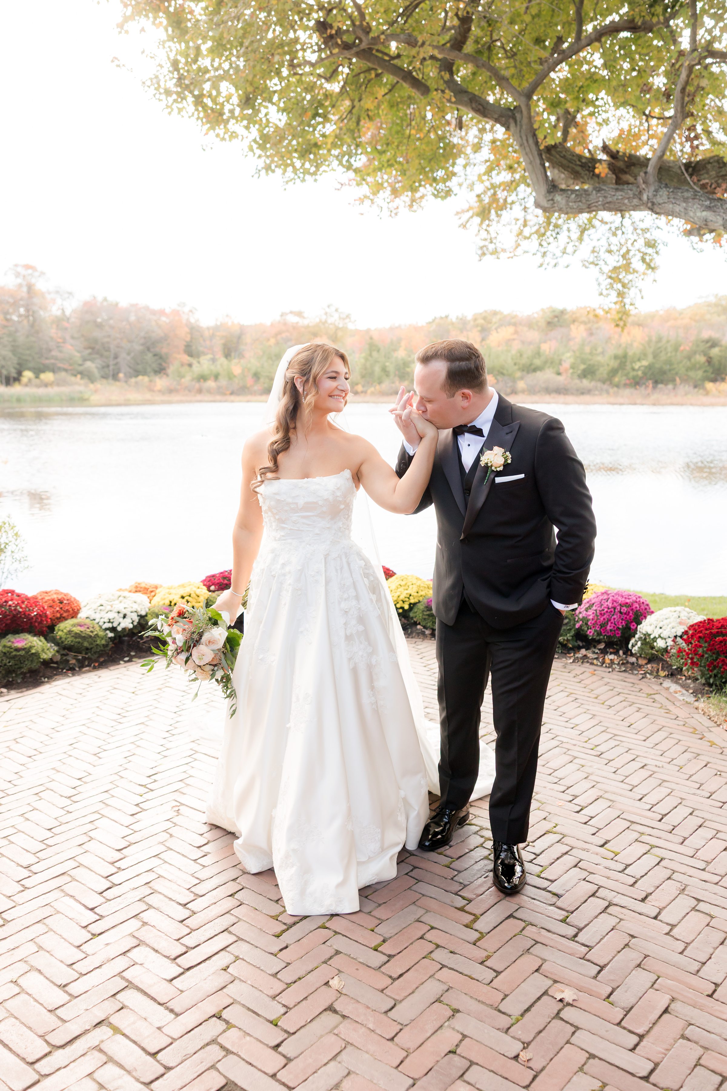 Groom kisses the bride’s hand while she smiles, holding her bouquet by the lakeside.