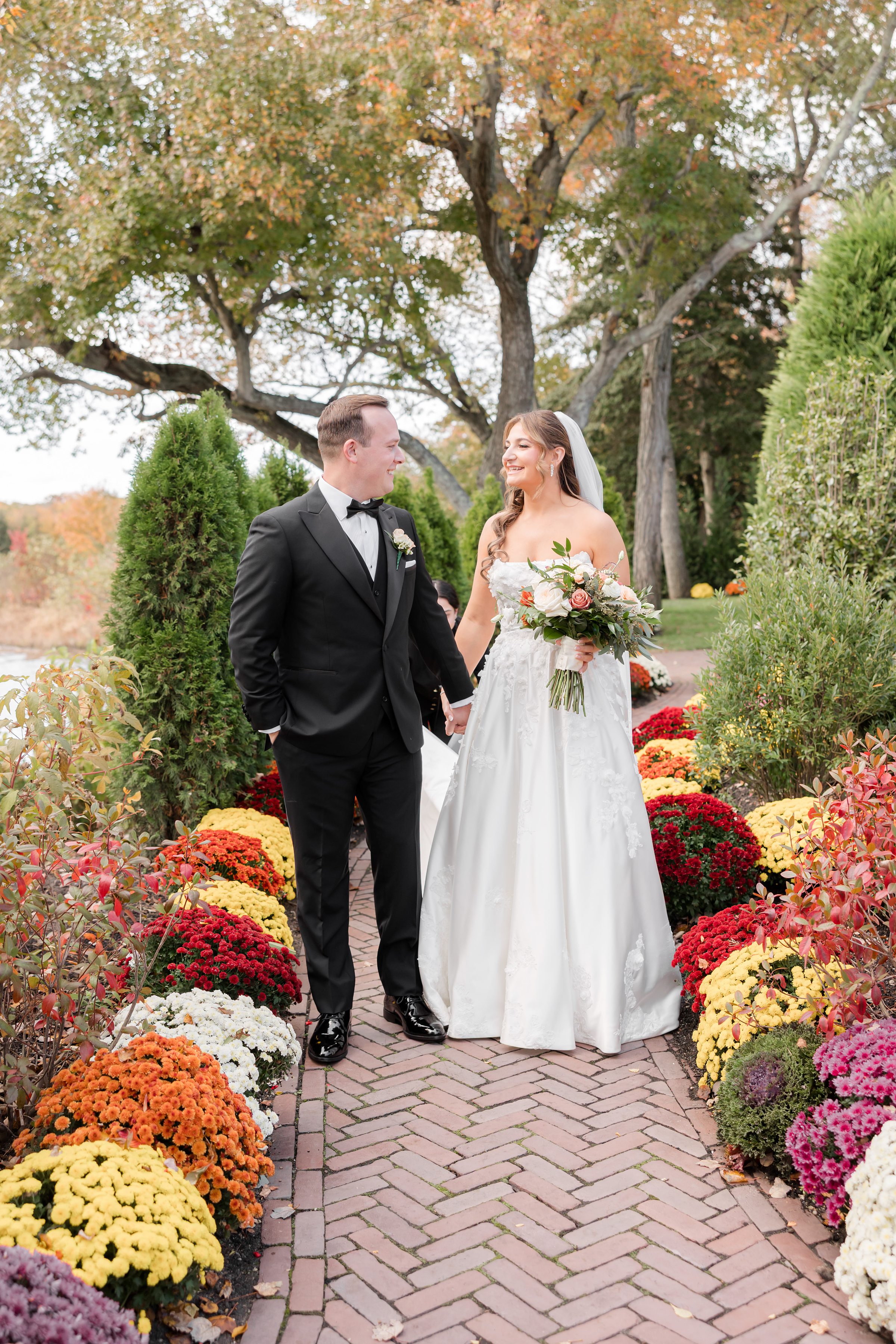Bride and groom walking together down a garden path lined with bright autumn flowers.