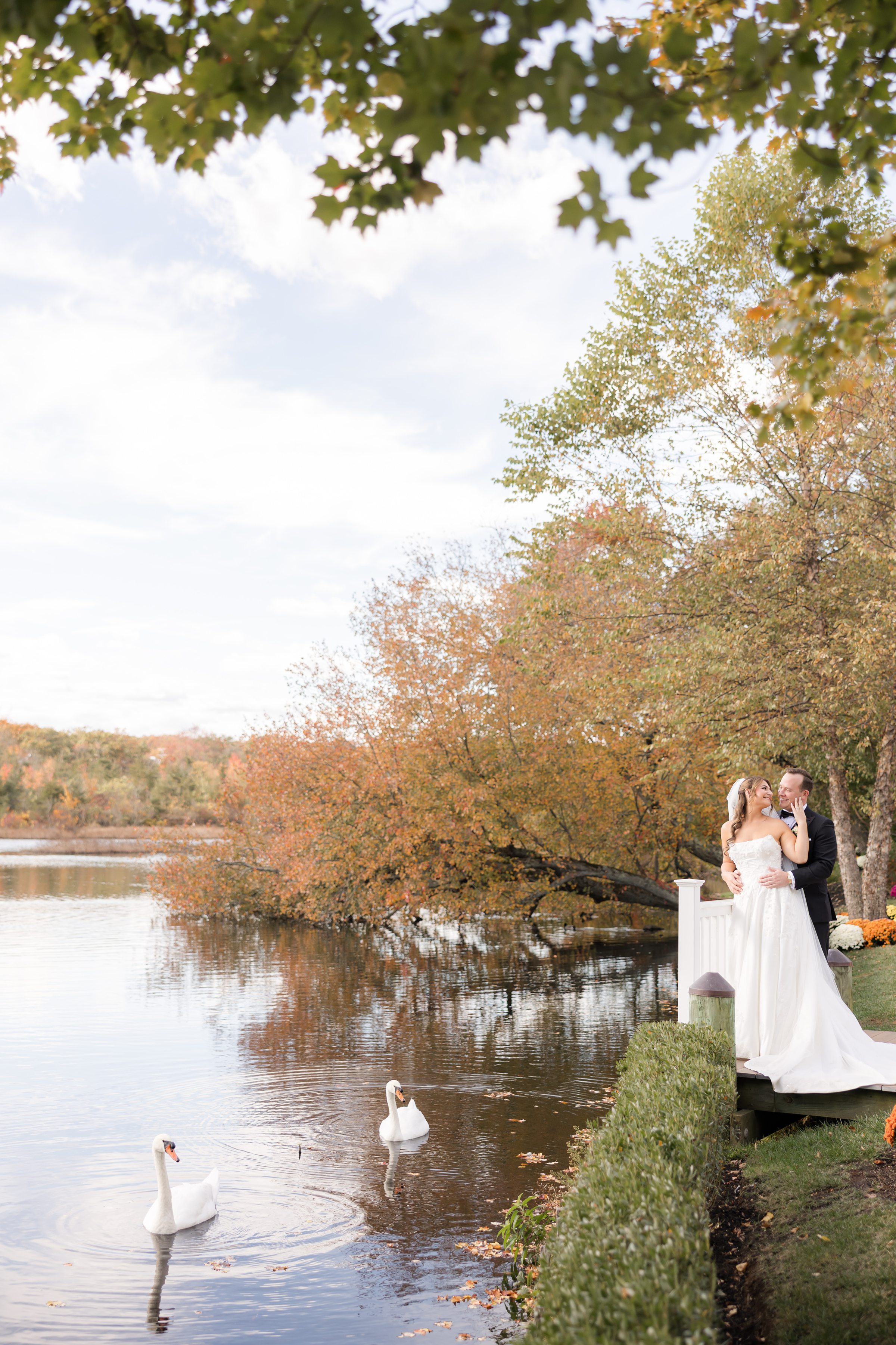 Bride and groom embrace by a peaceful lakeside, surrounded by glowing autumn trees, sharing a quiet, romantic moment beneath soft natural light.