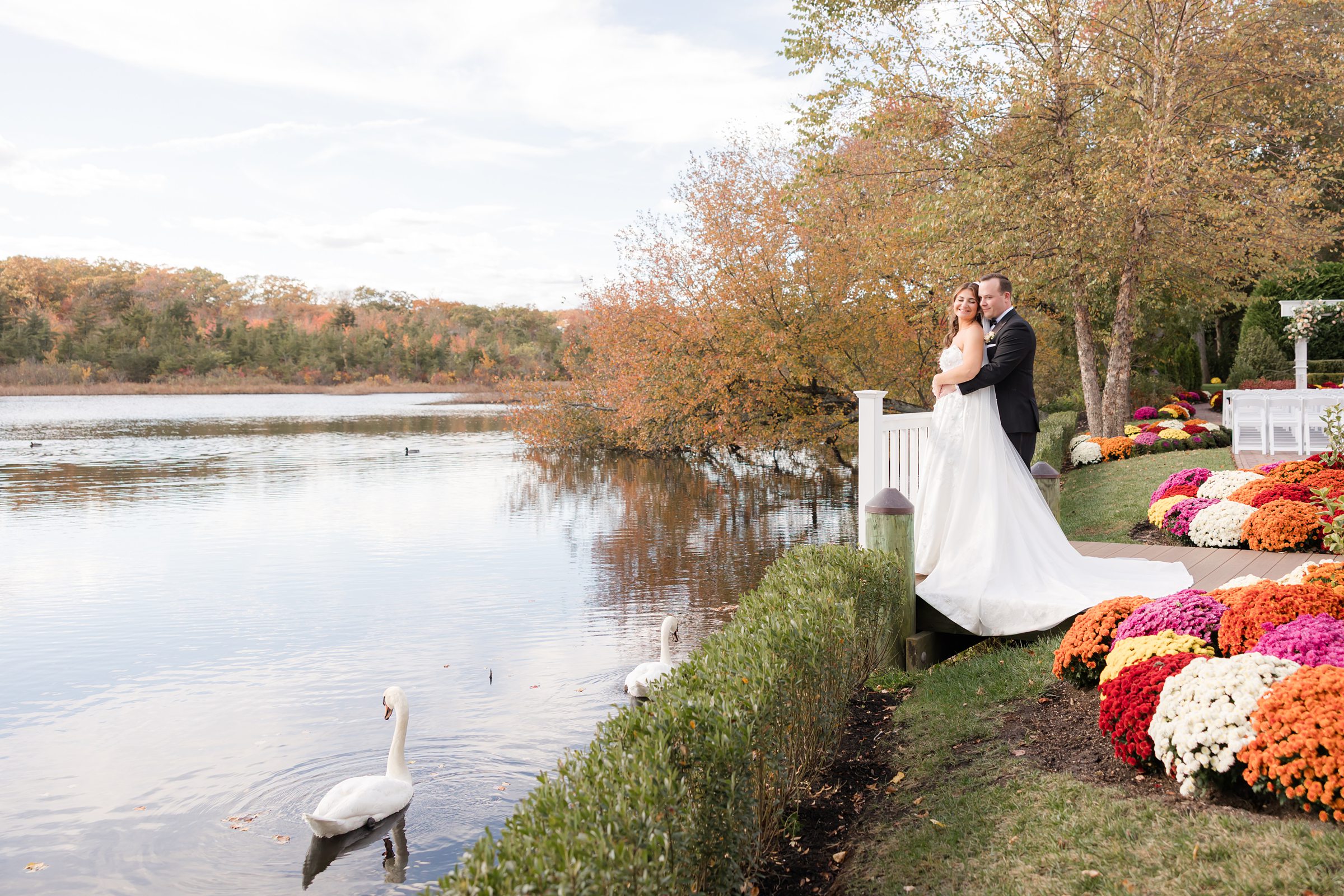 Bride and groom embracing on a small platform by a lakeside, with a swan gliding nearby and vibrant autumn flowers surrounding them.