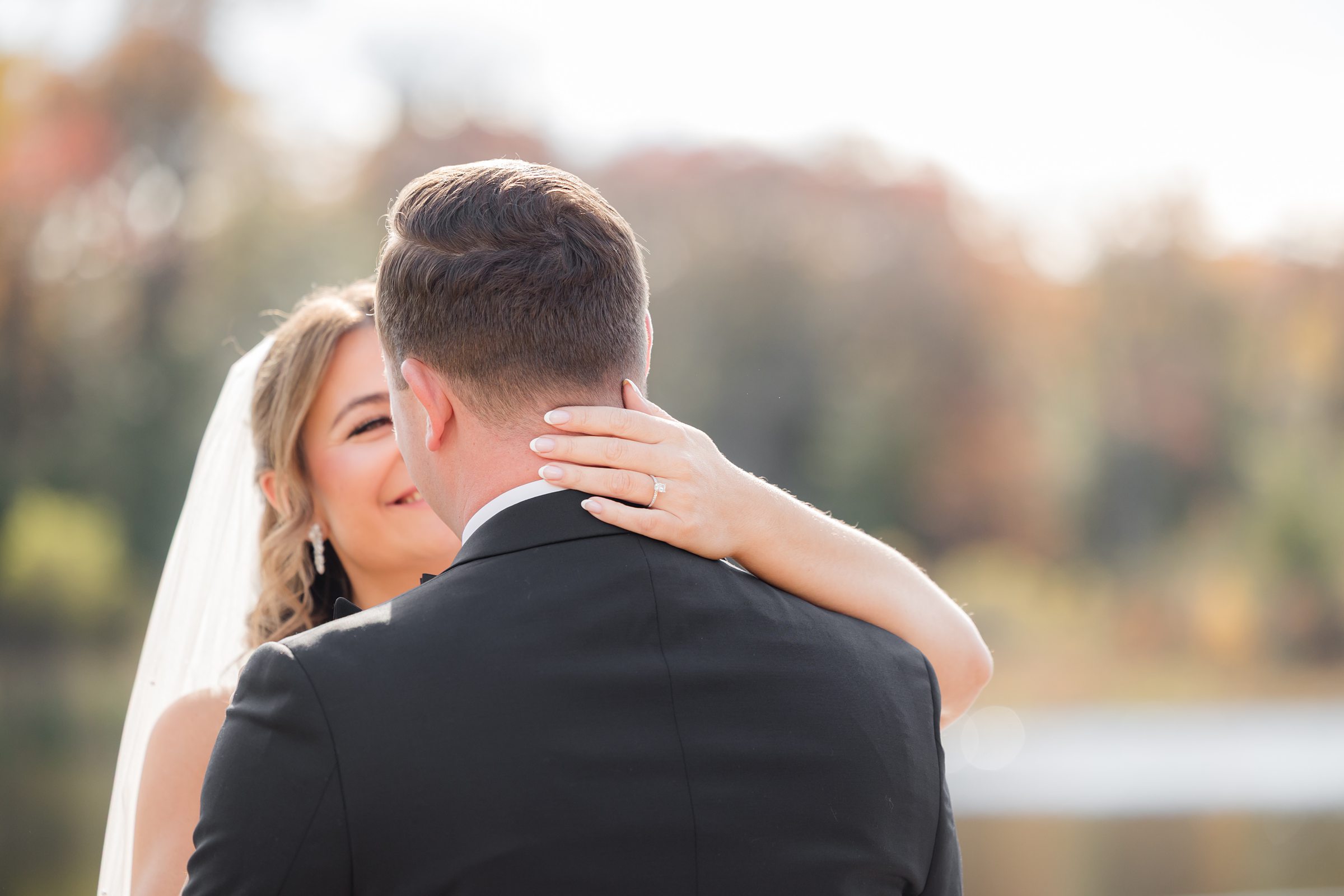 Bride and groom embrace closely outdoors, the bride smiles over the groom’s shoulder, showing her ring.
