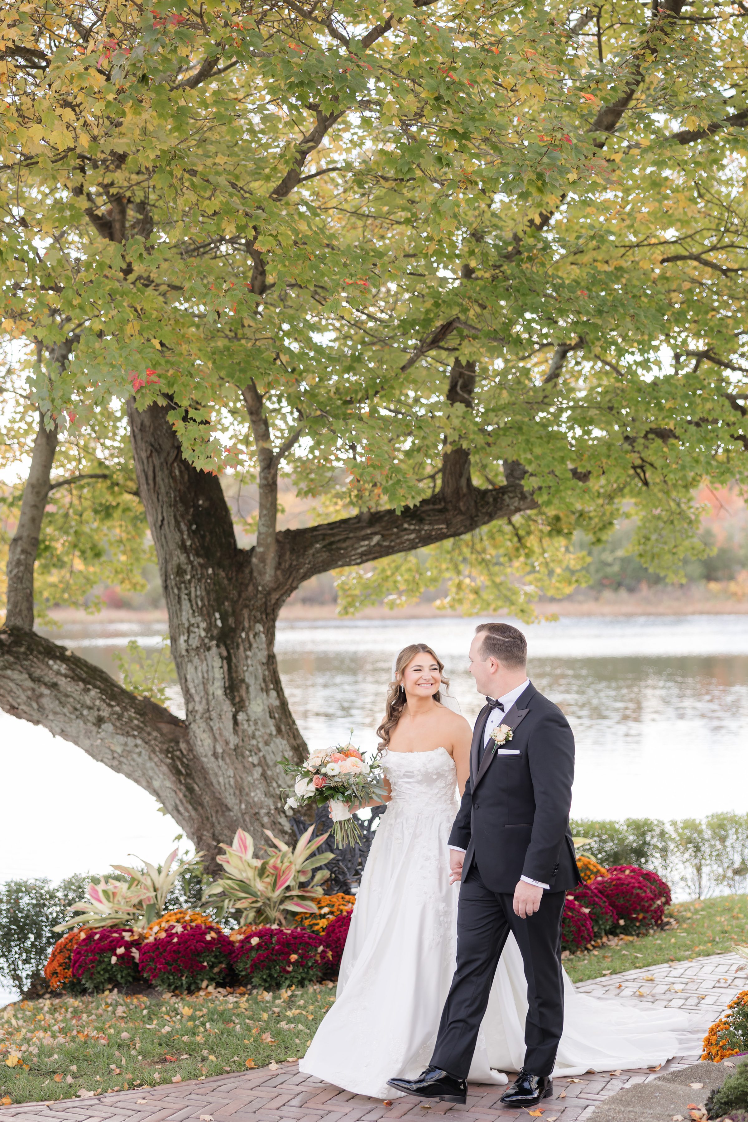 Bride and groom walking together under a large tree by the water, smiling at each other