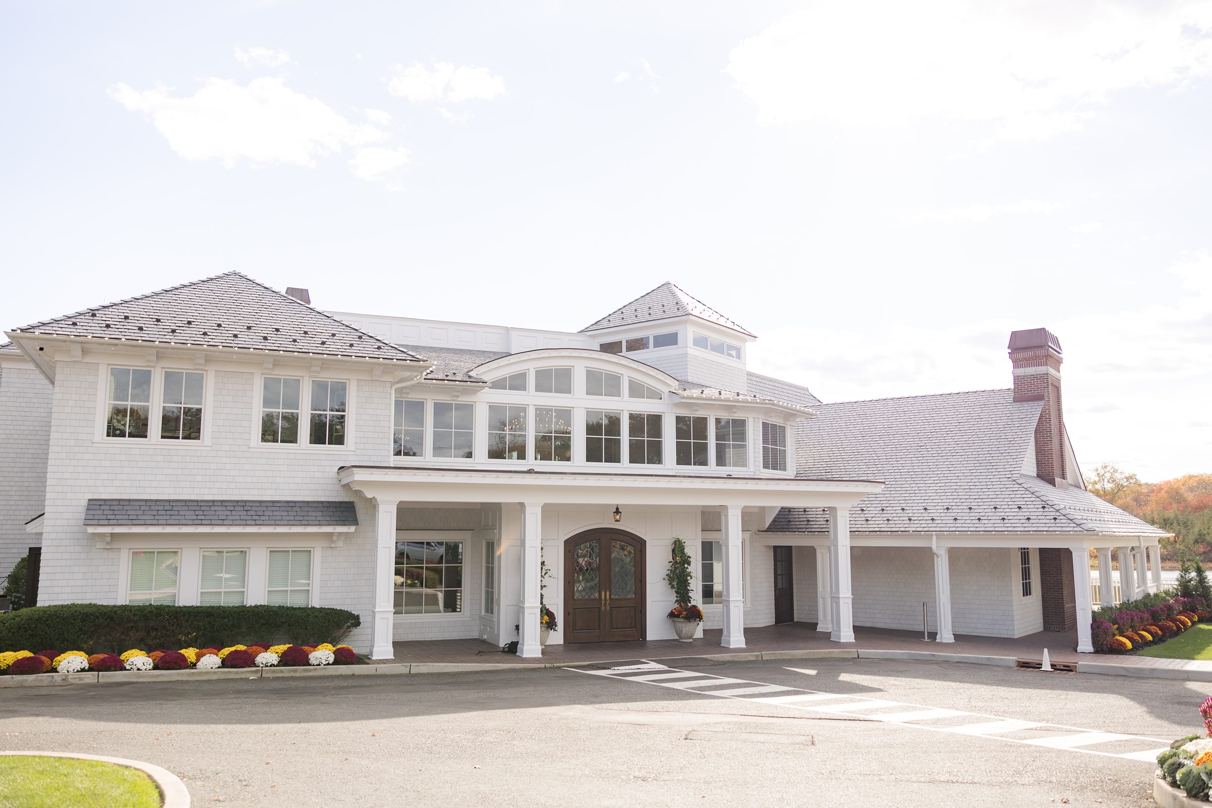 Wide exterior shot of a white wedding venue building with large windows and autumn landscaping.