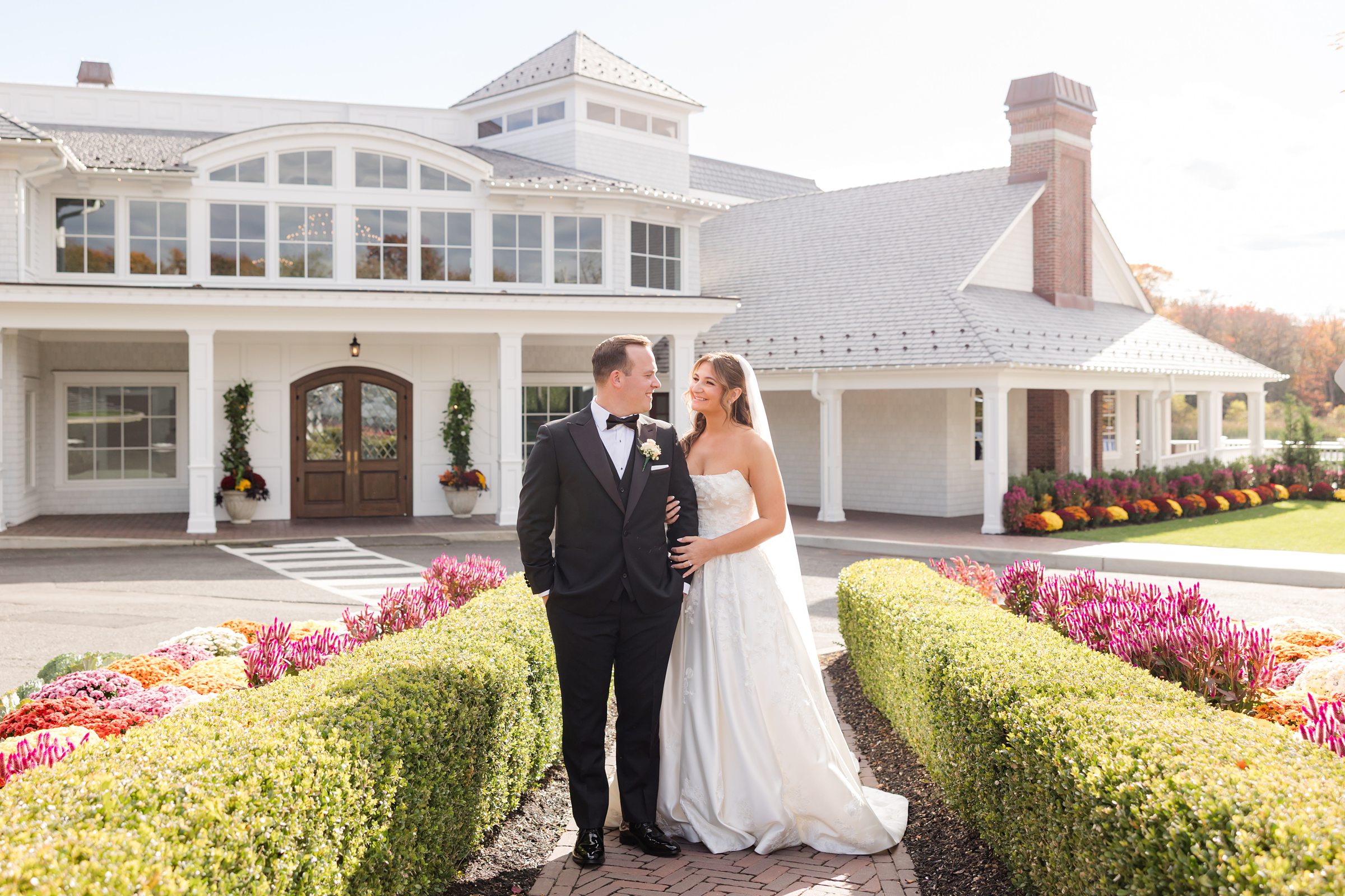 Bride and groom embracing in front of a white building, smiling at each other in soft sunlight.