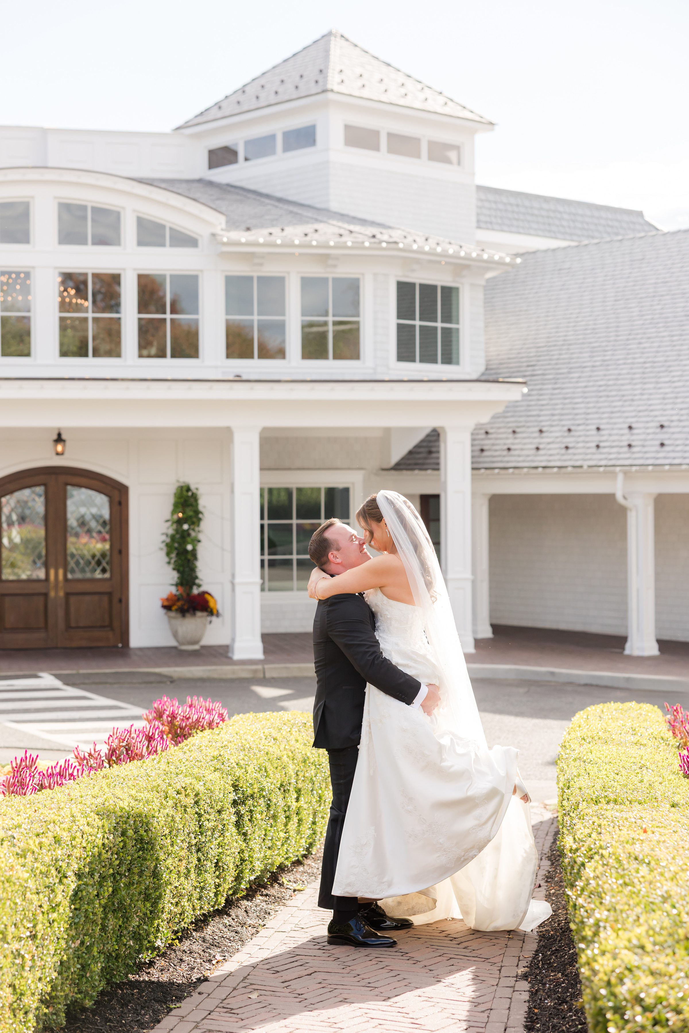 Bride and groom sharing a joyful embrace and kiss outside a bright white venue, surrounded by soft sunlight and autumn blooms.