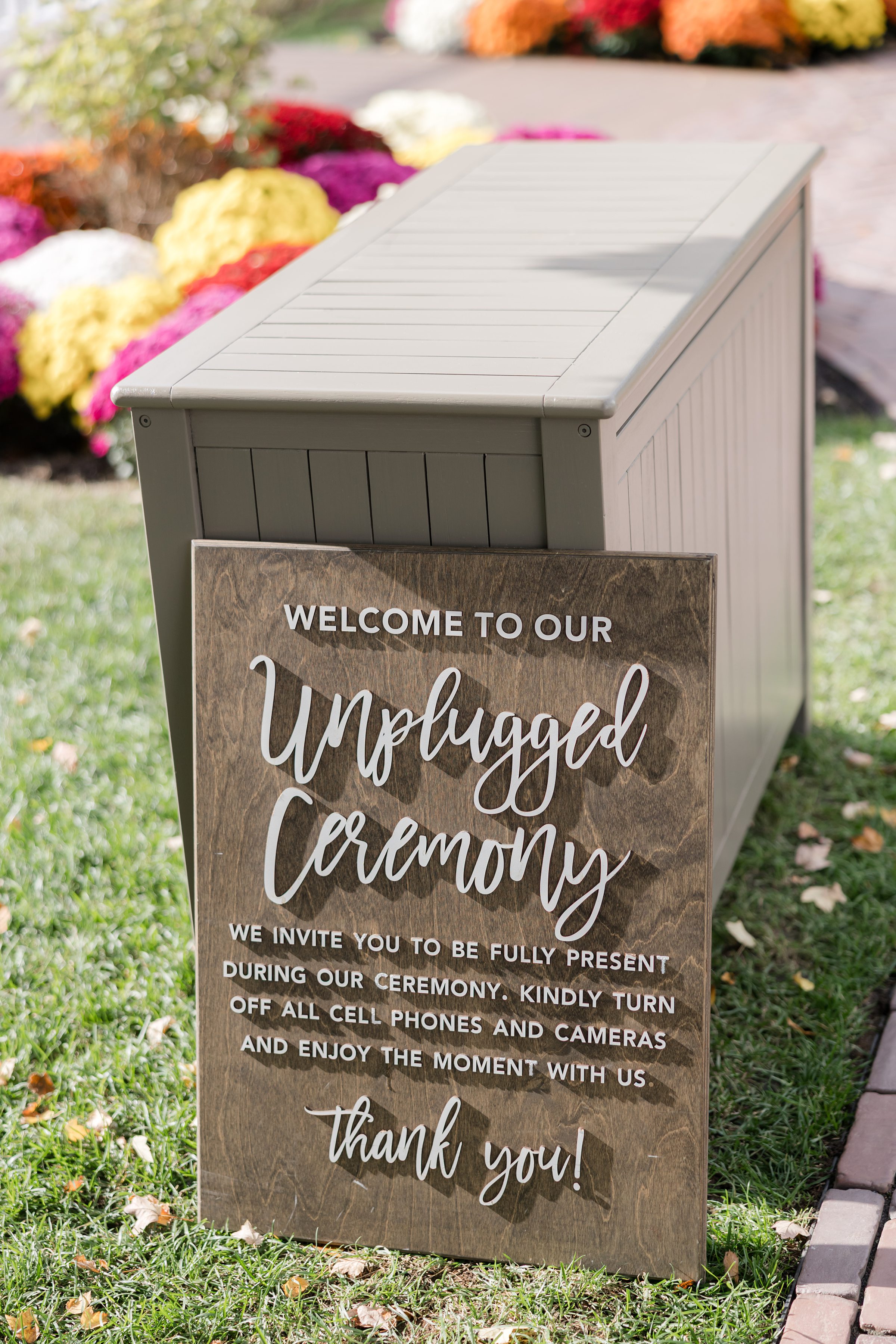 Wooden sign reading “Welcome to our unplugged ceremony” displayed on grass beside colorful autumn flowers, inviting guests to be fully present and put away devices.