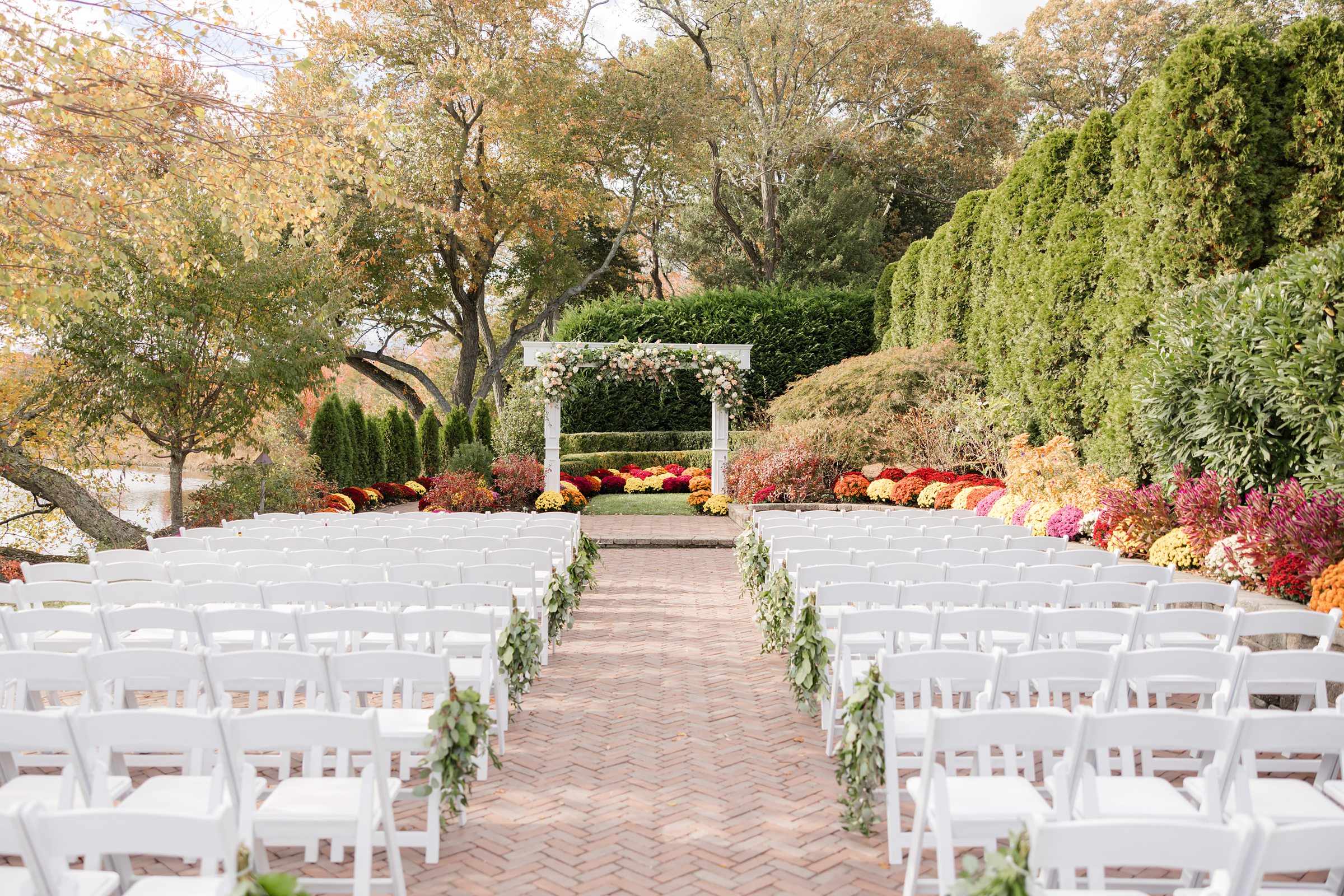 Outdoor wedding ceremony setup with white chairs lining a brick aisle, leading to a floral arch surrounded by lush greenery and vibrant autumn flowers.