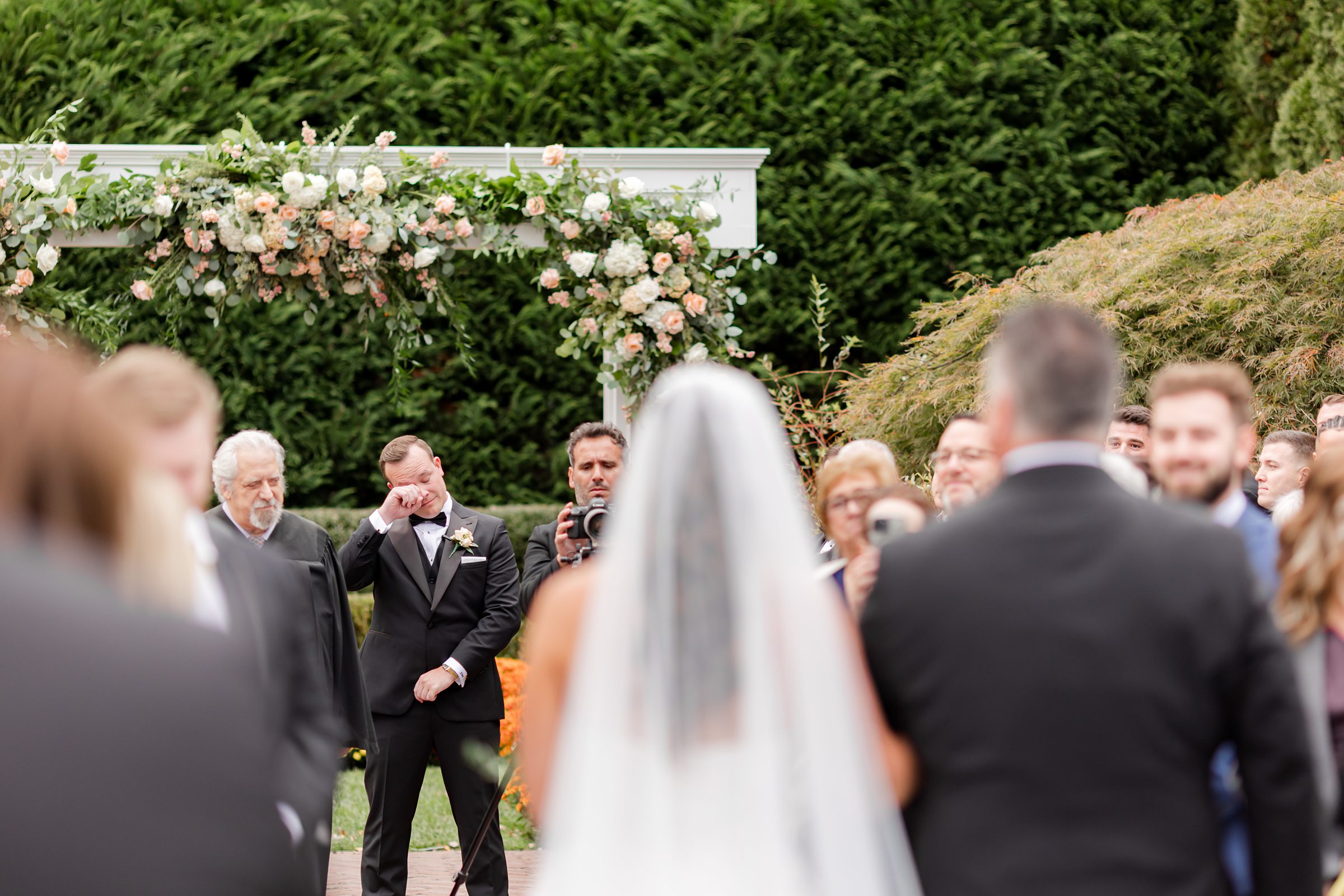 Bride walking down the aisle with her father toward the ceremony arch, as the groom waits emotionally in the background surrounded by guests.