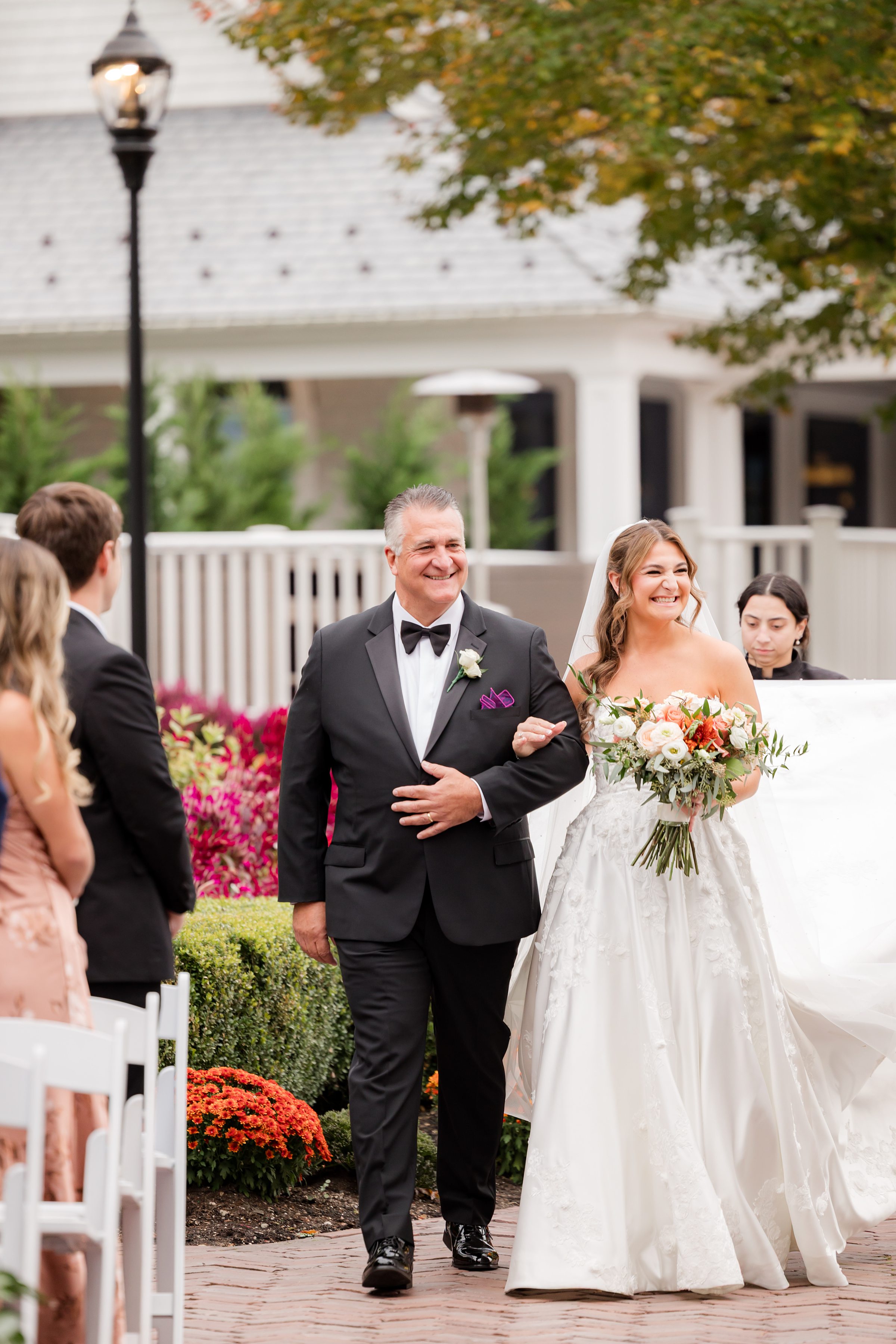 Bride walking down the aisle with her father, both smiling warmly as she carries a bouquet of soft, romantic blooms surrounded by guests and autumn florals.