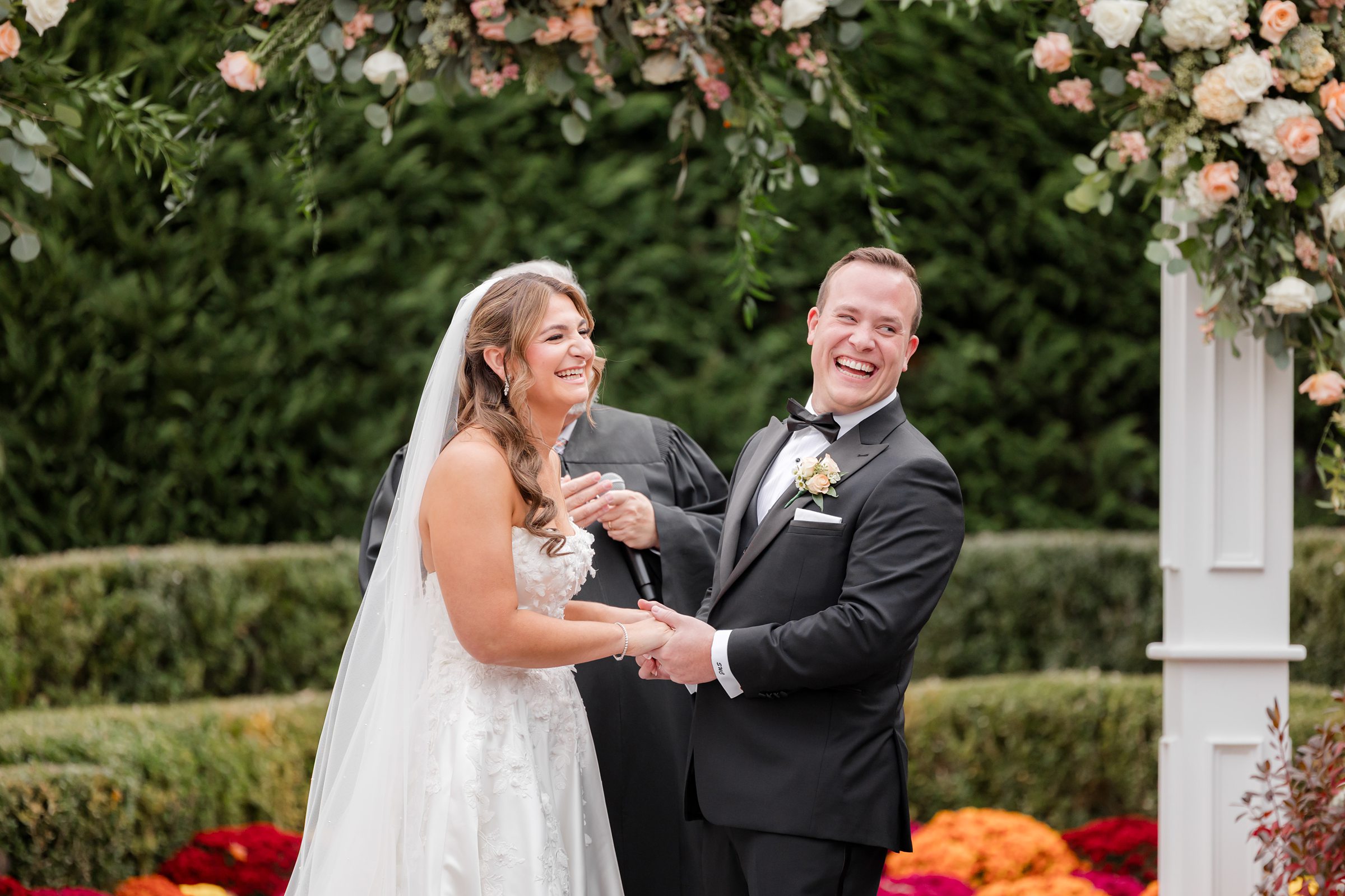 Bride and groom holding hands and smiling during the ceremony under a floral arch