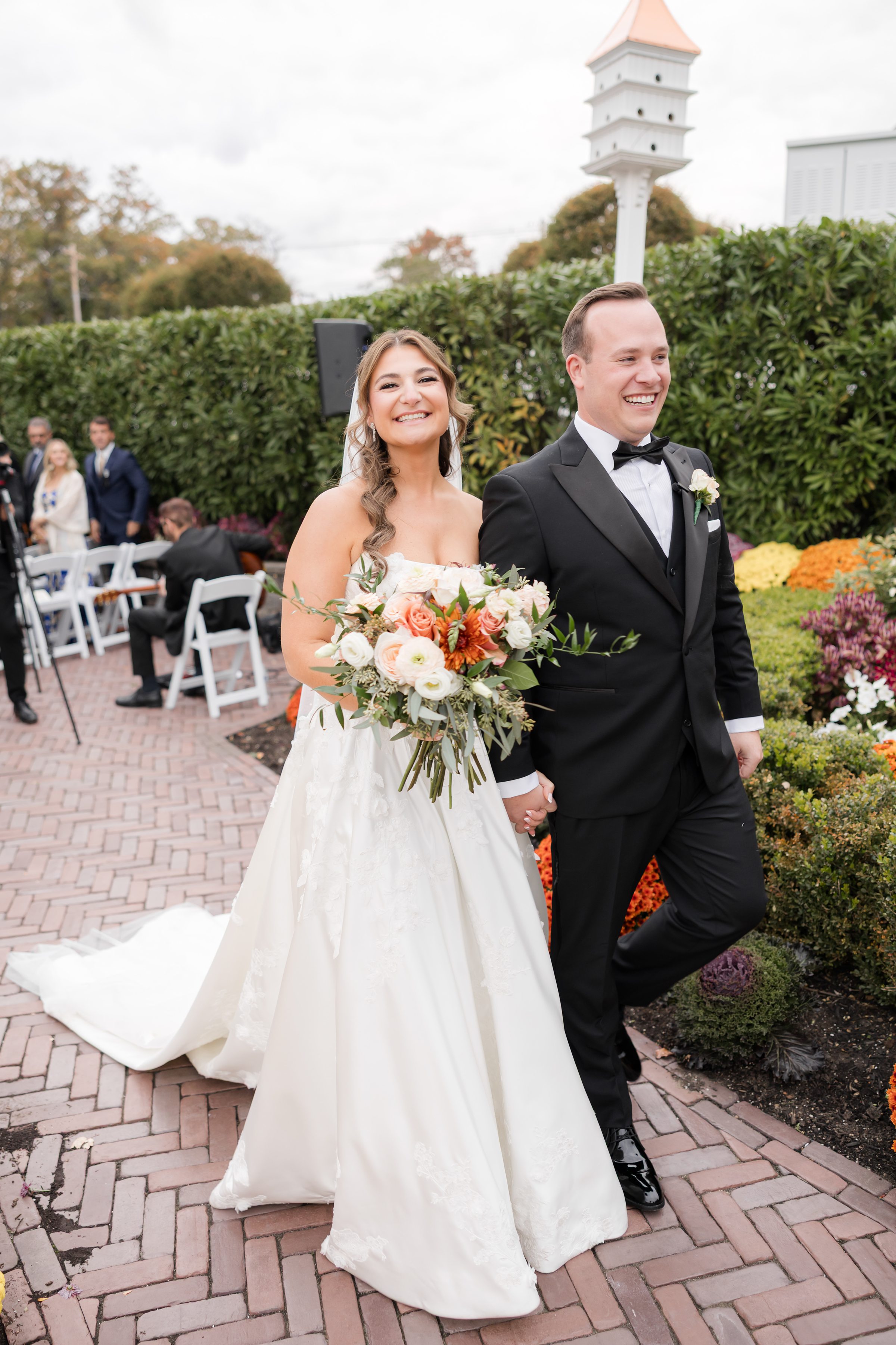 Newlyweds walking together down the garden path, smiling with joy as the bride holds a lush bouquet and guests look on among colorful autumn flowers.