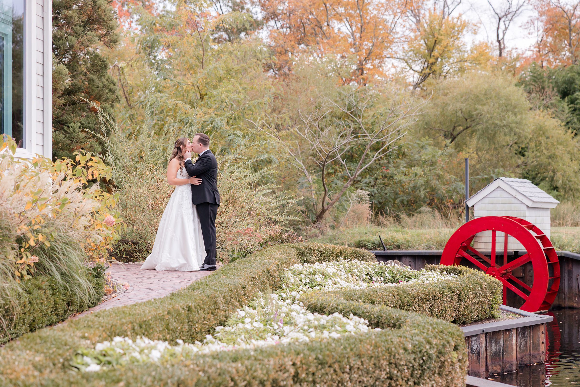 Bride and groom sharing a romantic kiss along a garden path by the water, surrounded by soft greenery, autumn foliage, and a charming red water wheel.