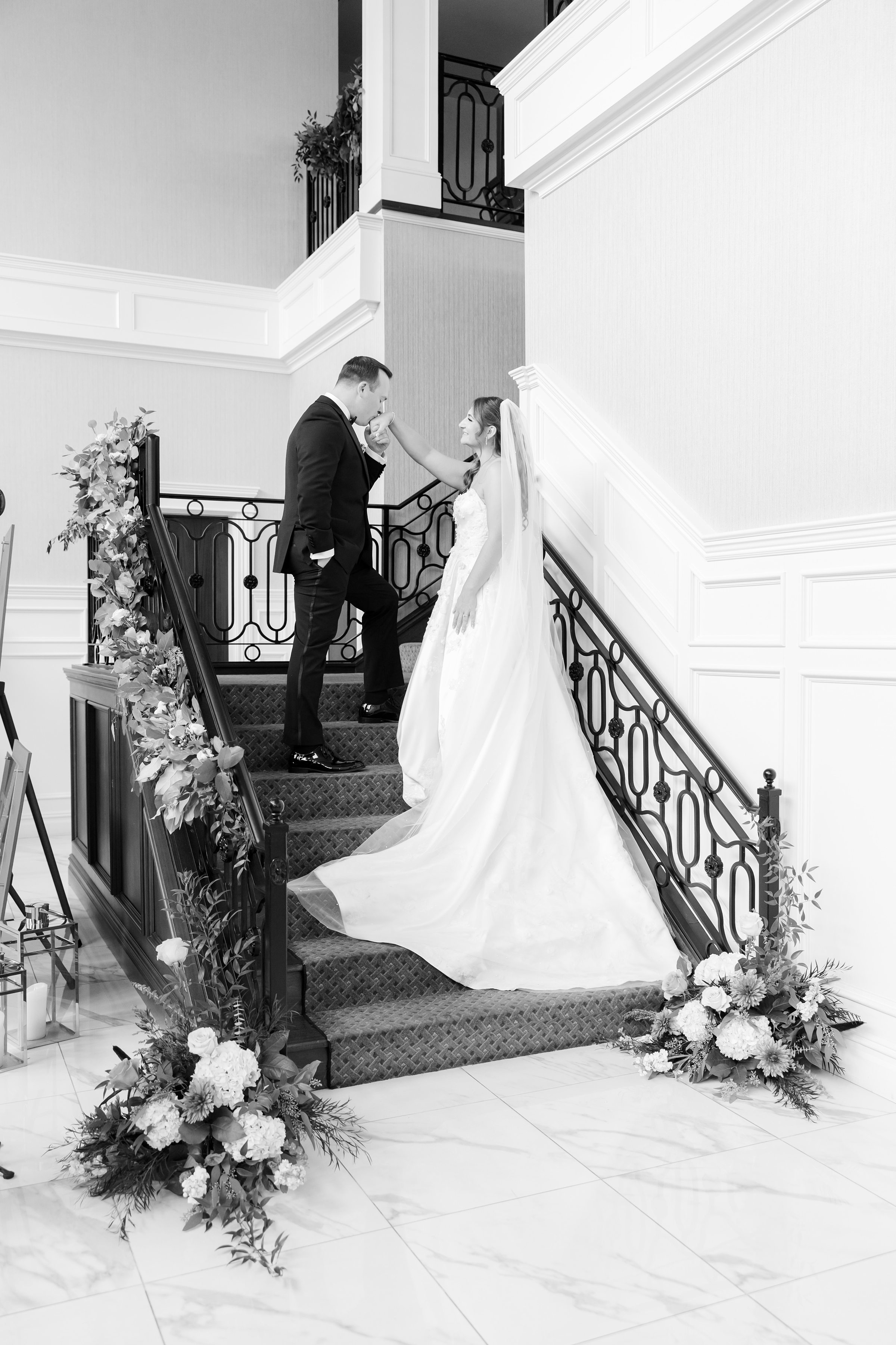 Groom kissing bride’s hand as she stands on a staircase in her gown.