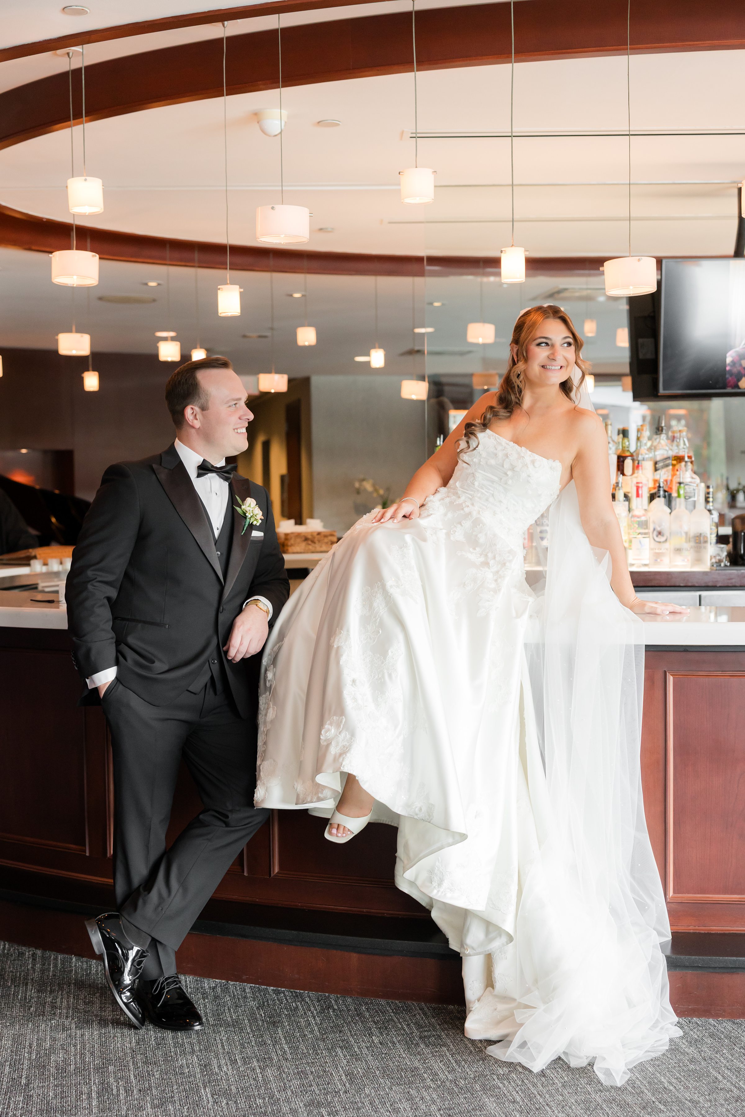 Bride sitting on a bar counter while groom leans beside her in a playful pose.