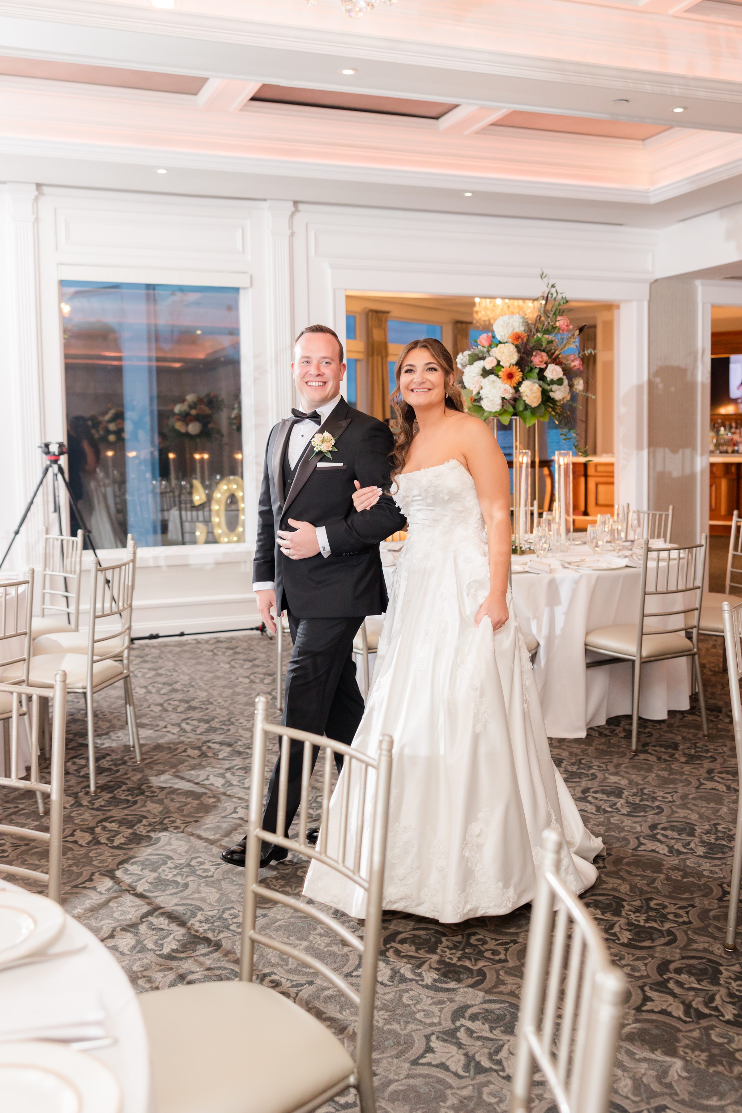 Bride and groom walking together into the reception space, smiling, with elegantly set tables and floral centerpieces in the background.