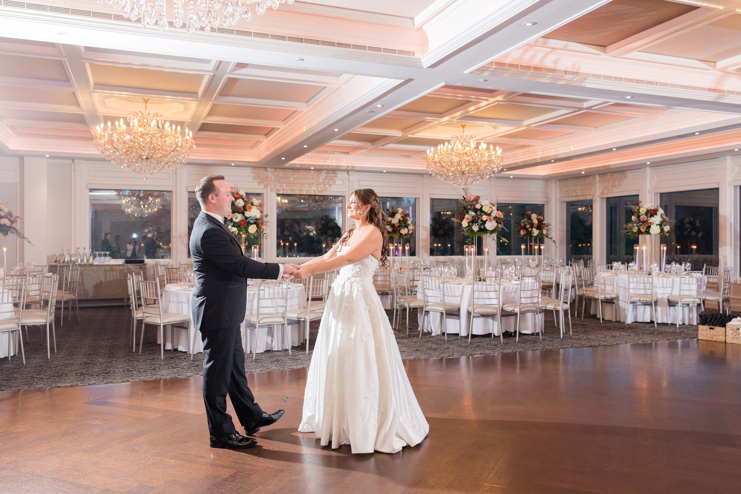 Bride and groom holding hands and smiling at each other on the dance floor in an elegant reception hall with chandeliers and round tables in the background.