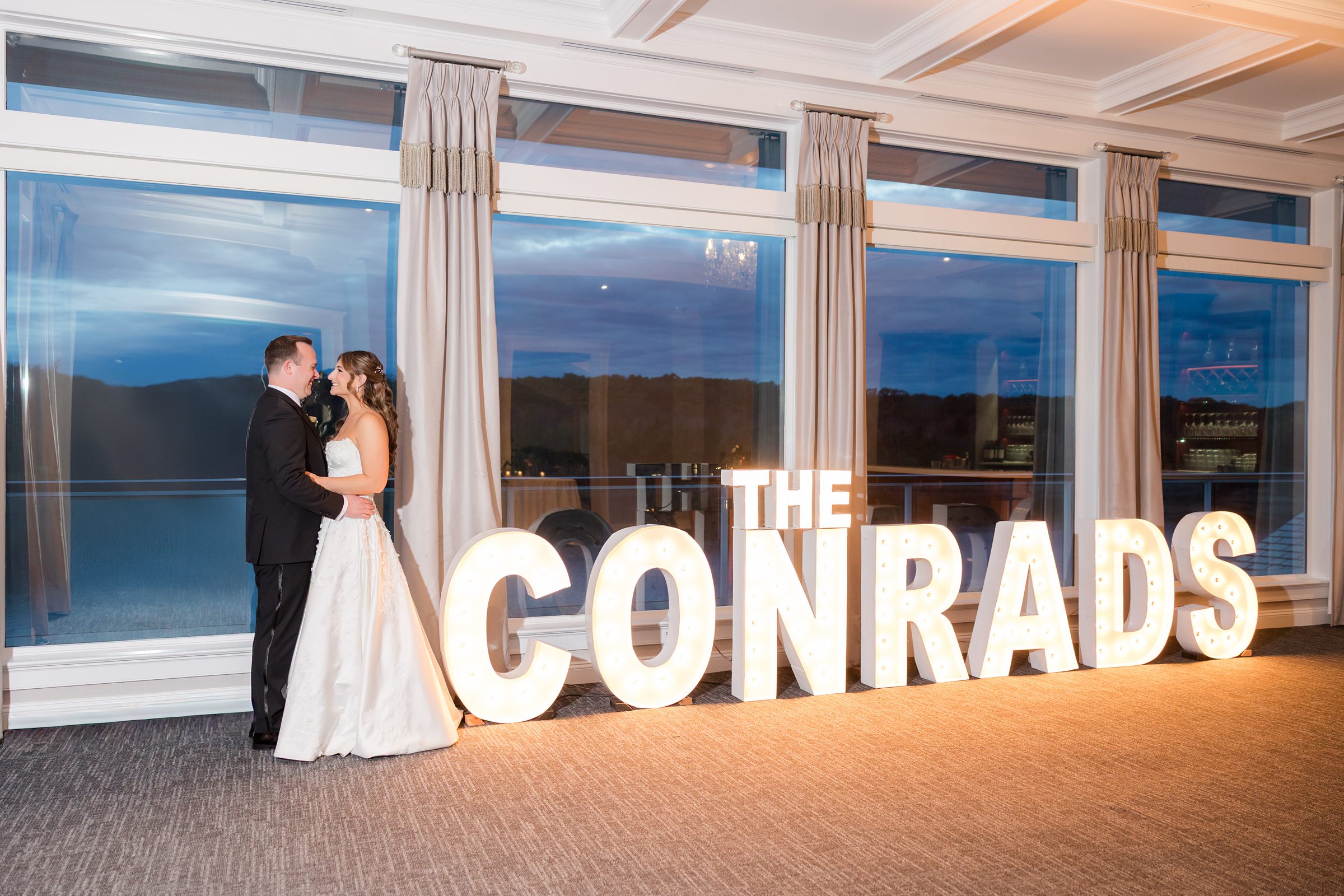 Bride and groom embracing beside illuminated “THE CONRADS” sign at sunset