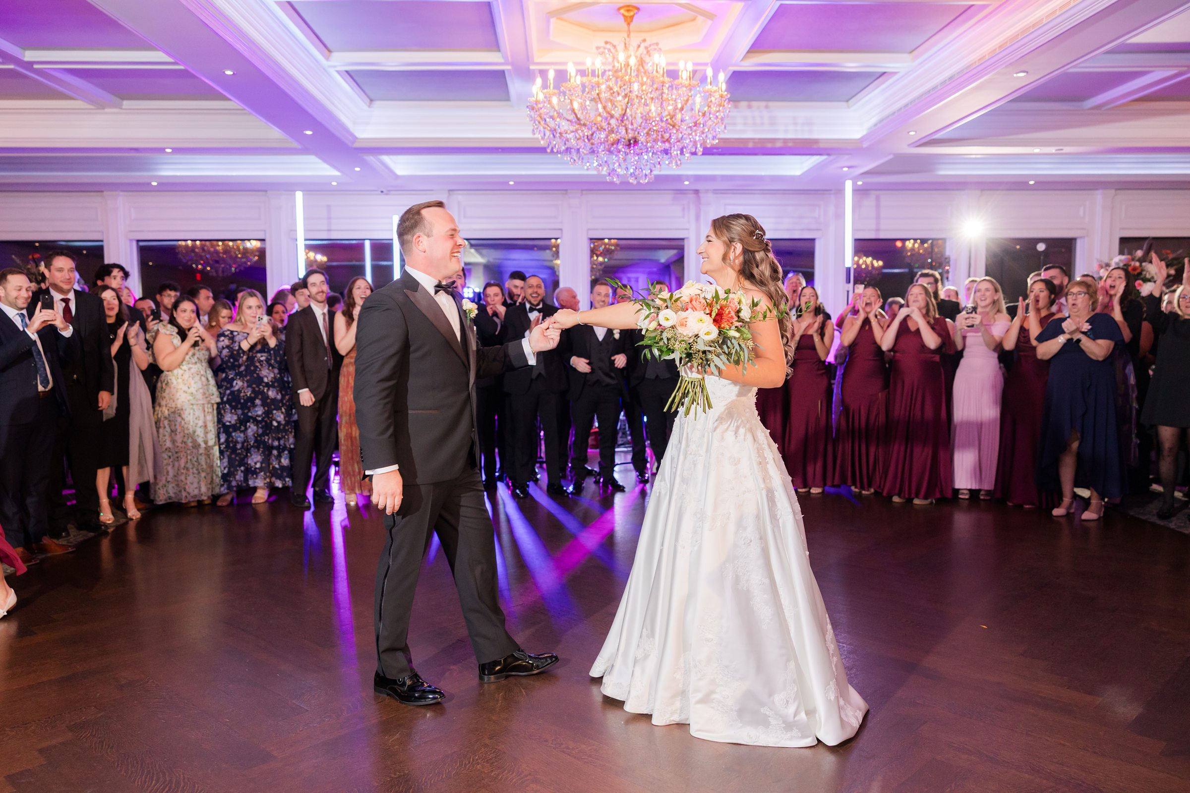 Bride and groom holding hands and smiling during their dance under chandeliers