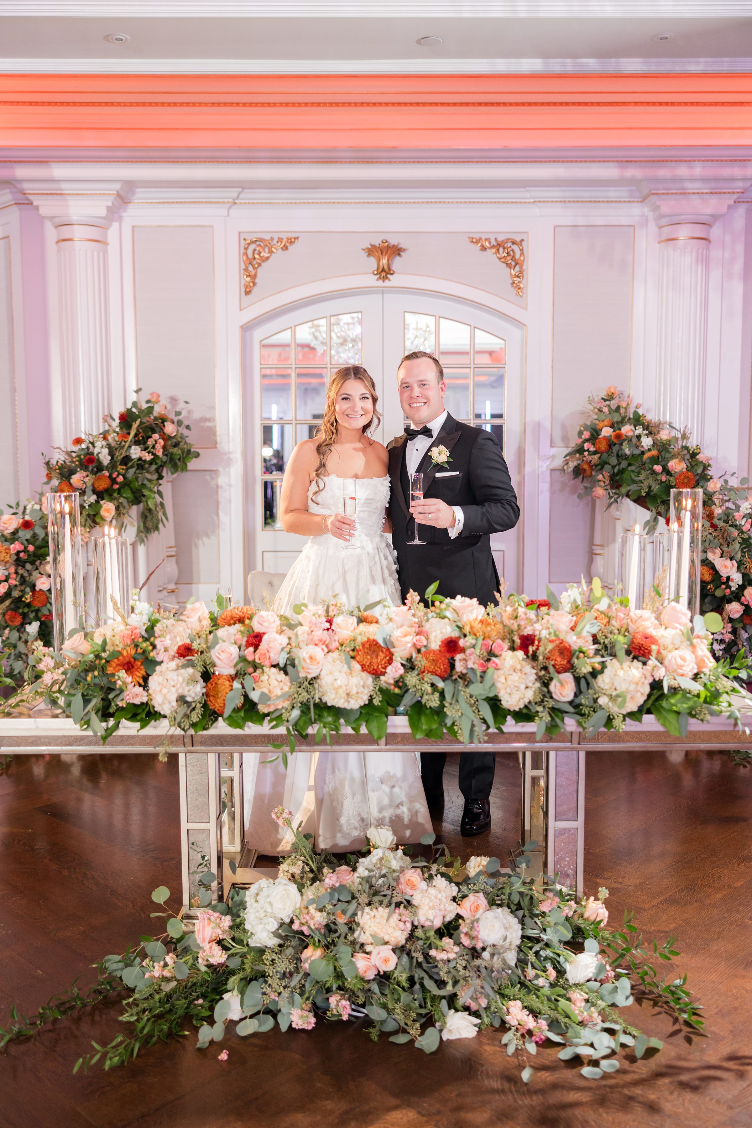 Bride and groom standing behind a long head table filled with lush floral arrangements.