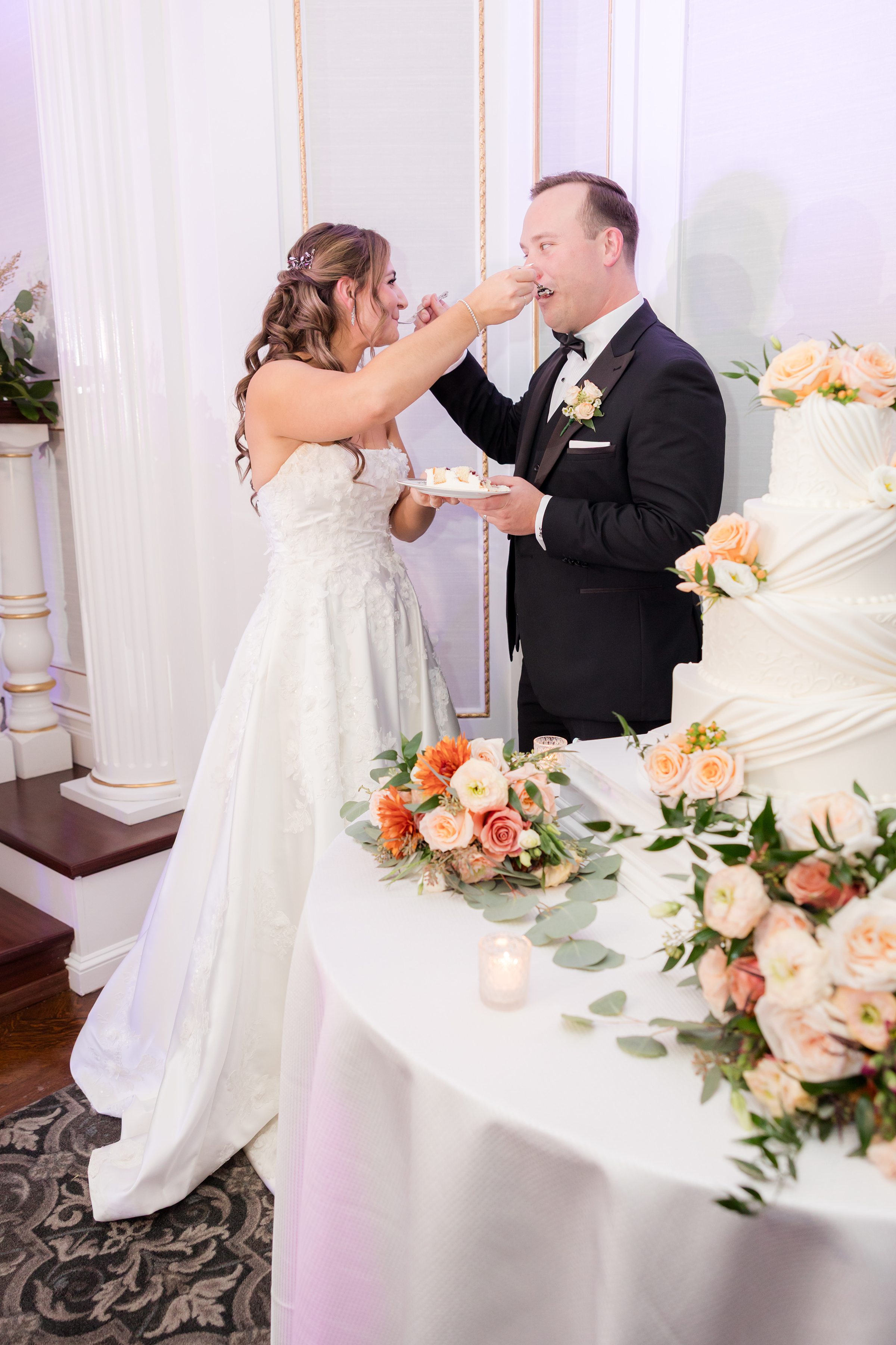 Bride feeding groom a bite of wedding cake beside a tiered cake decorated with peach roses and greenery on a floral-adorned table.