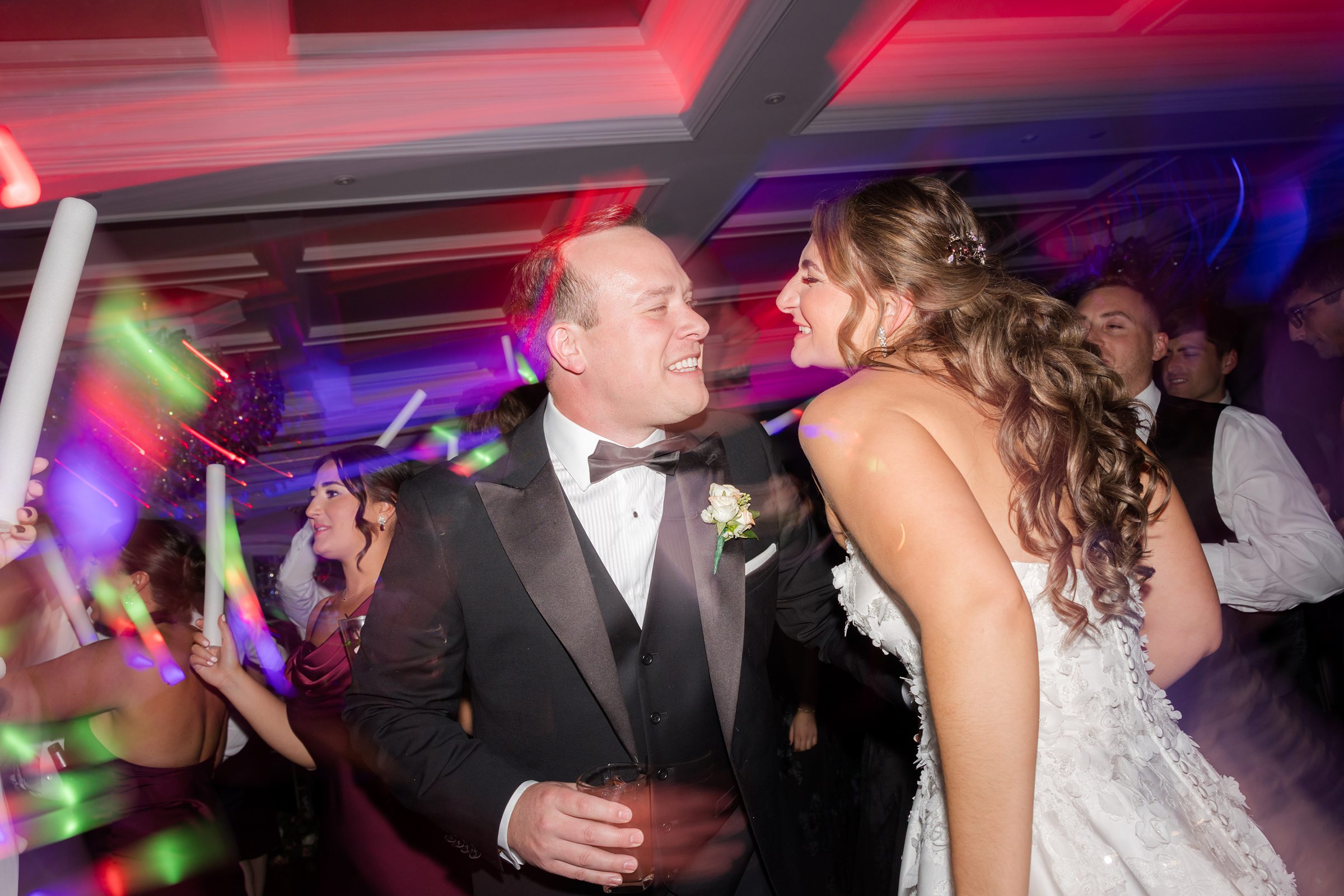 Bride and groom laughing together on a crowded dance floor with colorful lights.