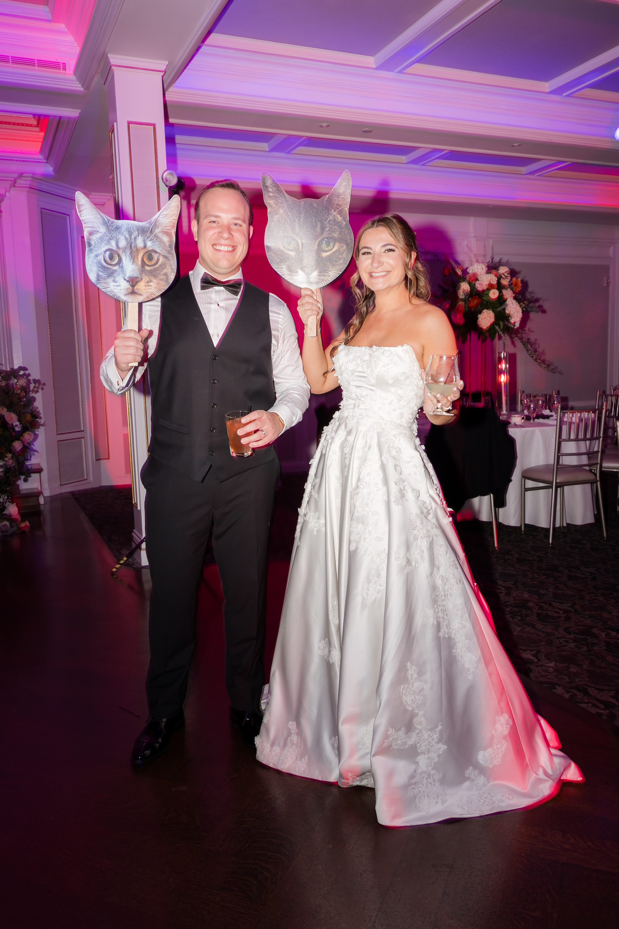 Bride and groom smiling at reception, holding drinks and playful cat-face props.