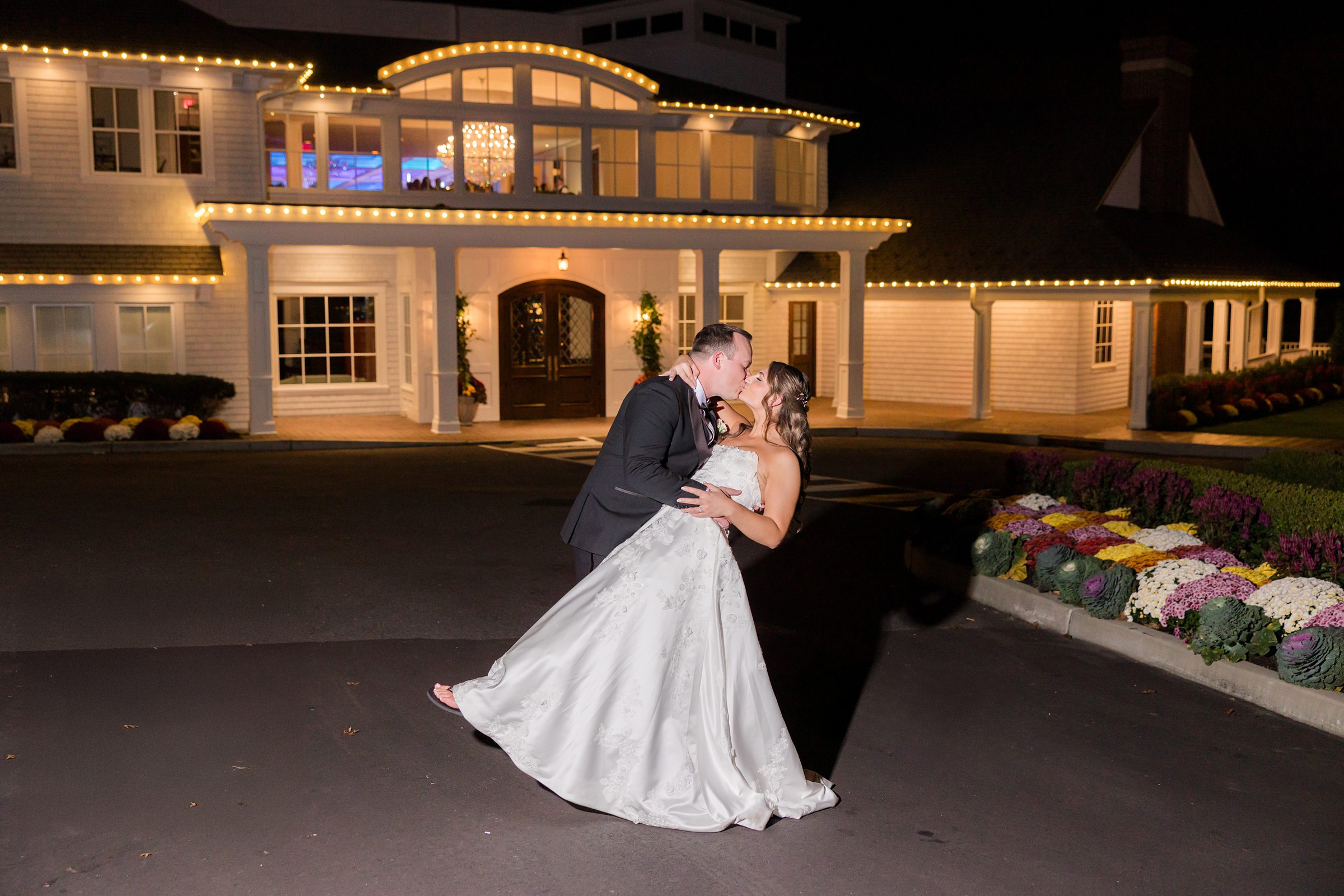 Bride and groom share a kiss in a dramatic dip pose outside a warmly lit wedding venue at night.
