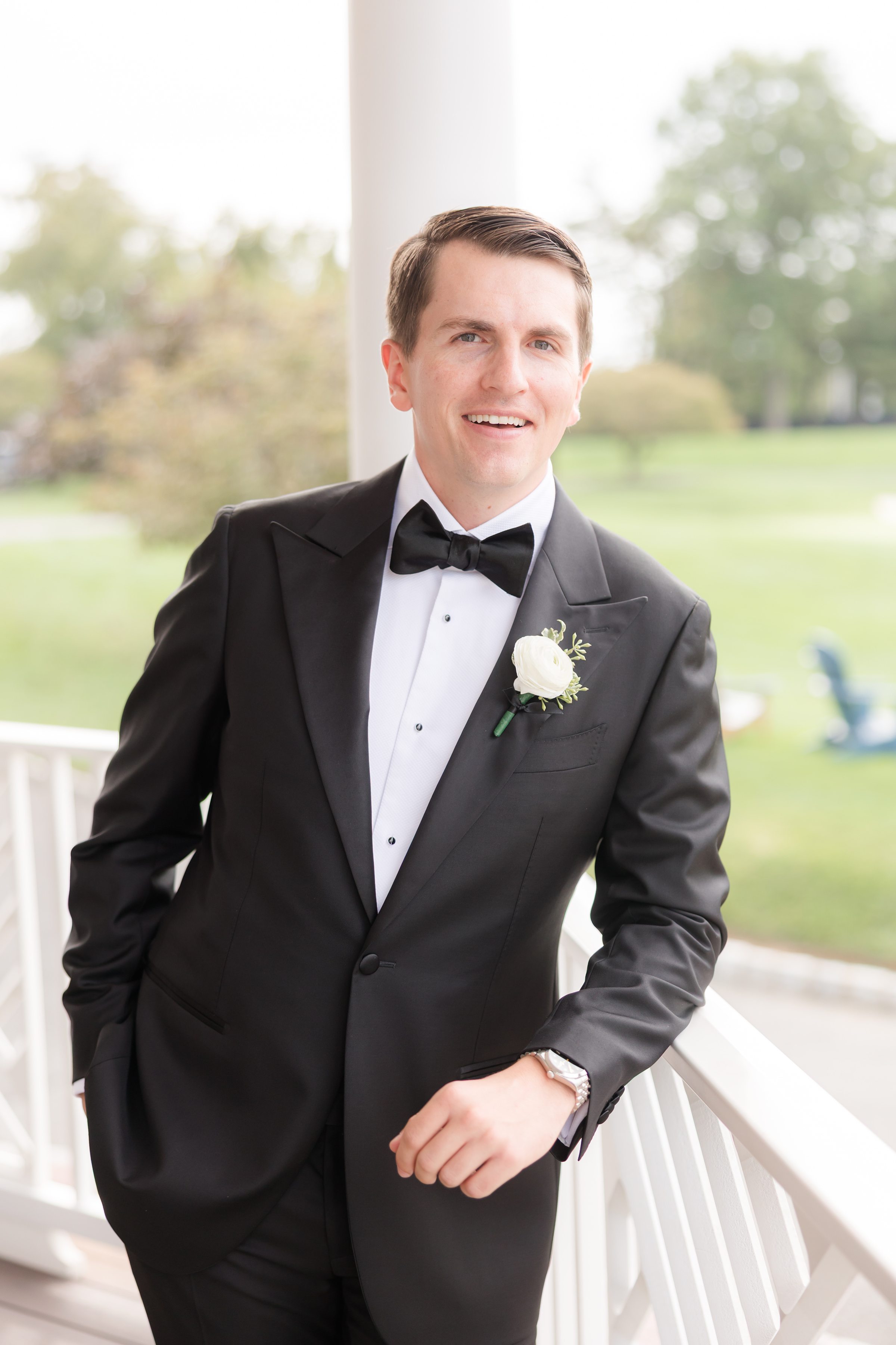 Groom leans casually on a porch railing, smiling warmly in his classic black tuxedo as he awaits the ceremony.