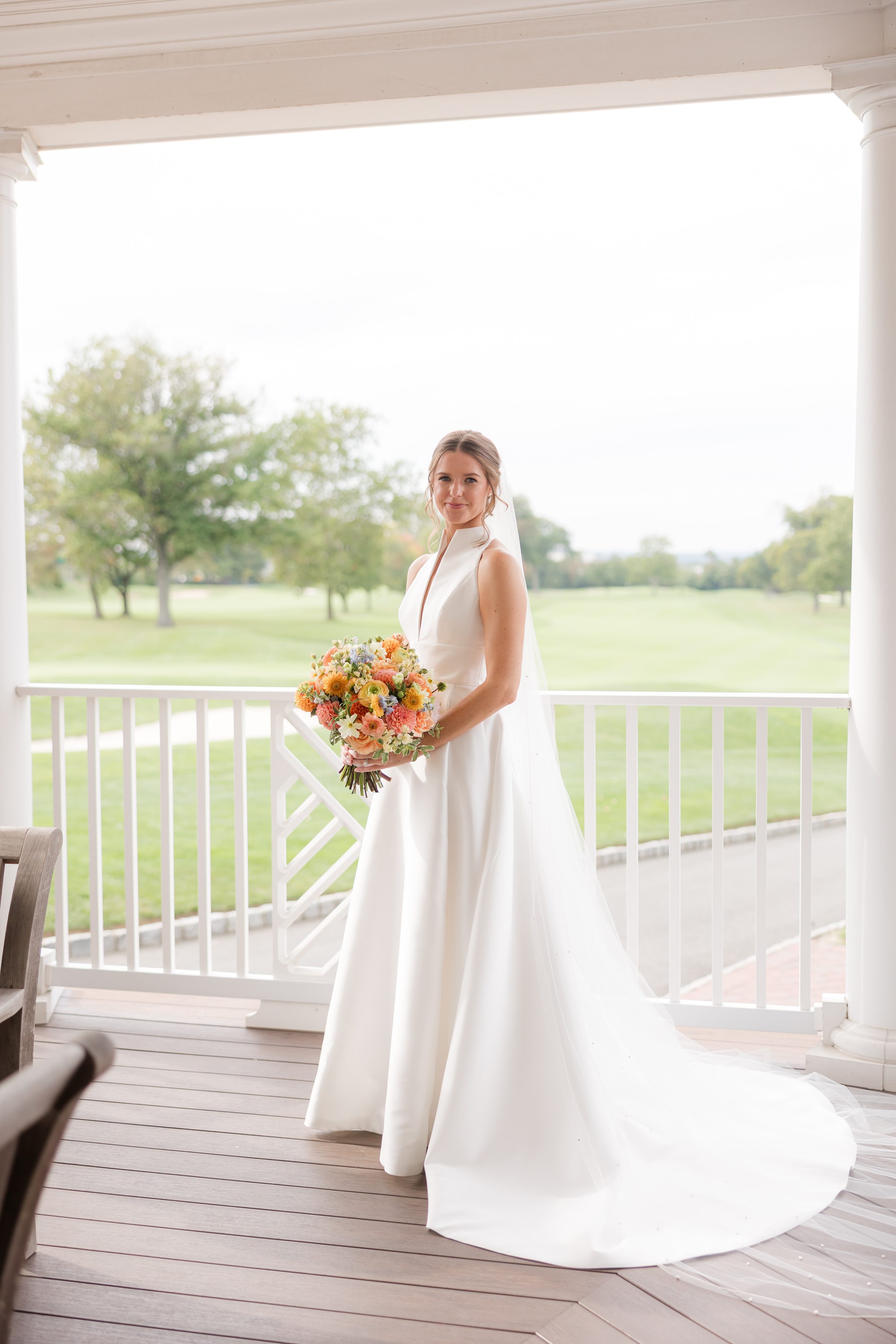 Bride stands gracefully on a sunlit porch, holding a vibrant bouquet, her expression serene and radiant.