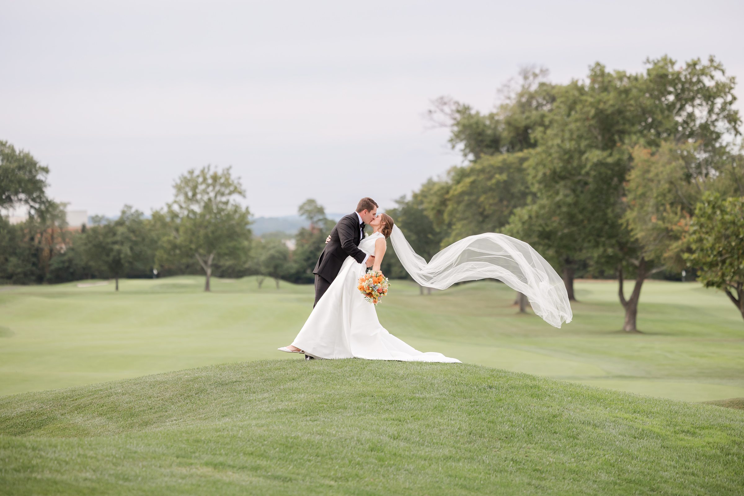 Bride and groom share a sweeping kiss atop a grassy hill, her veil drifting in the breeze like a soft, romantic whisper.