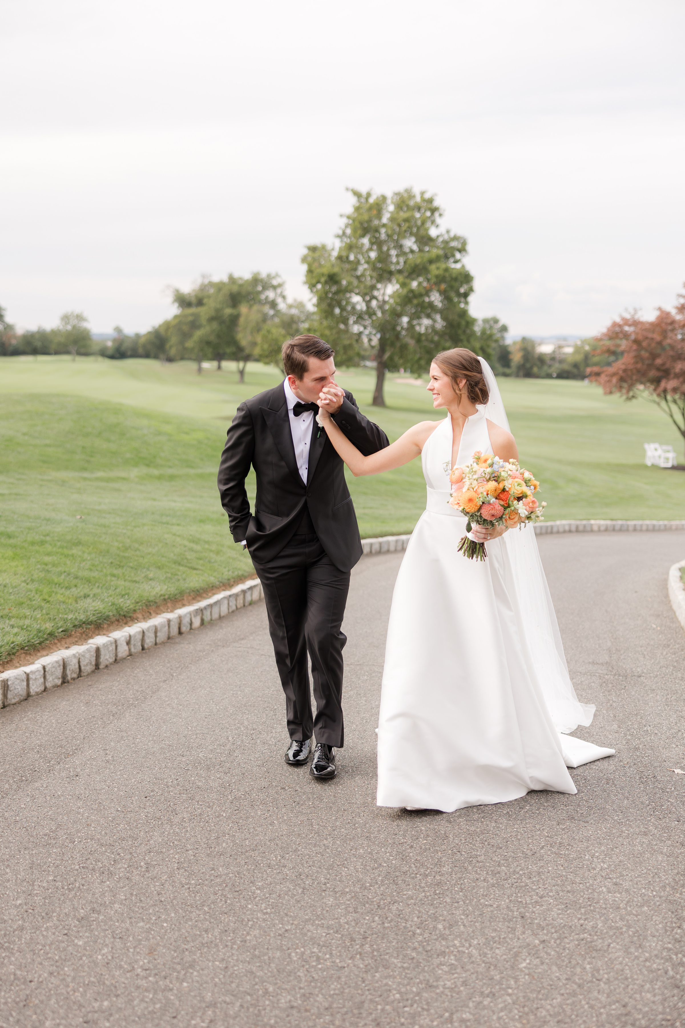 Groom lifts his bride’s hand for a gentle kiss as they walk together, sharing a moment of pure romance and devotion.