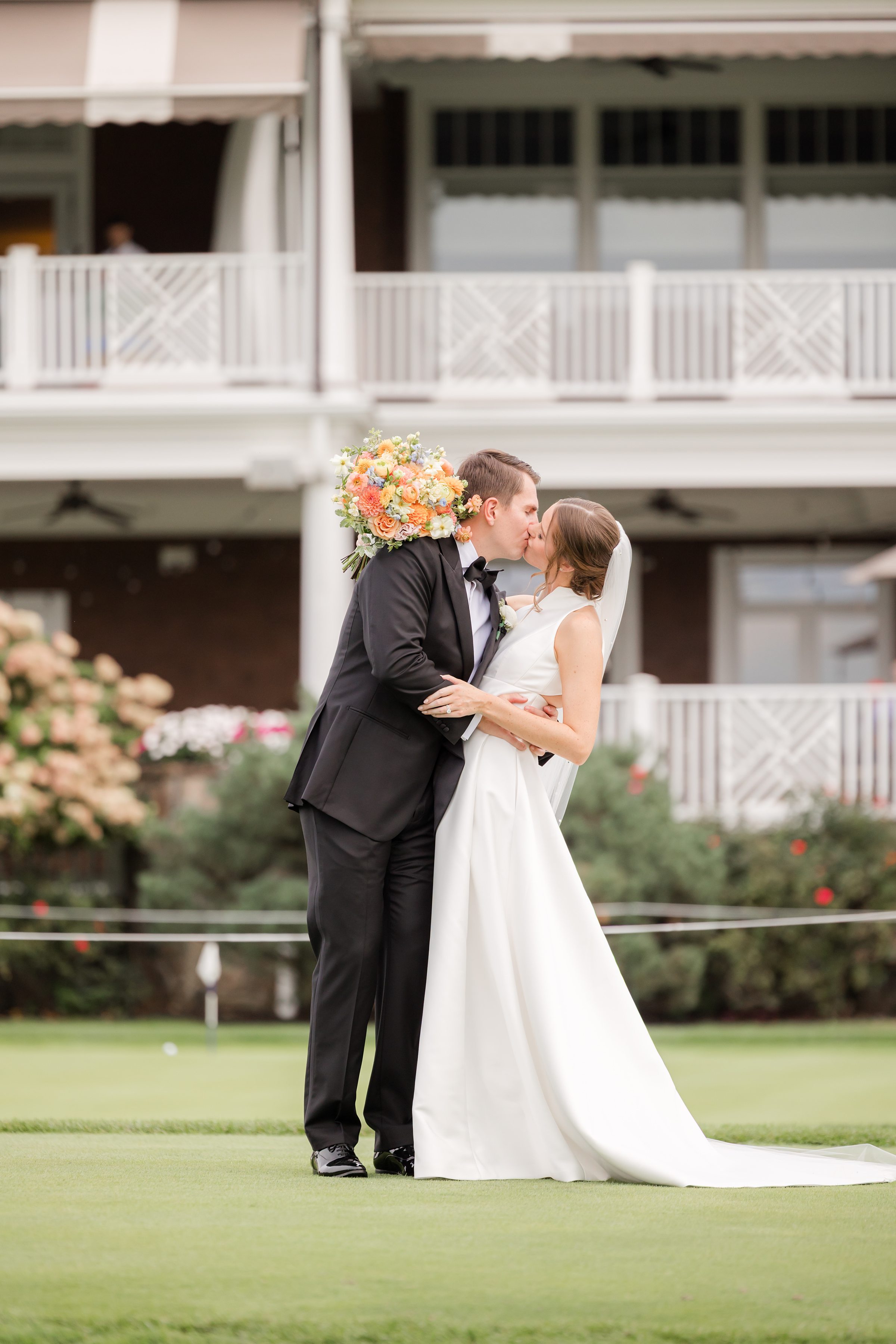 Outside a charming venue, the groom gently dips his bride into a kiss, her bouquet nestled between them as they savor a quiet, romantic moment.