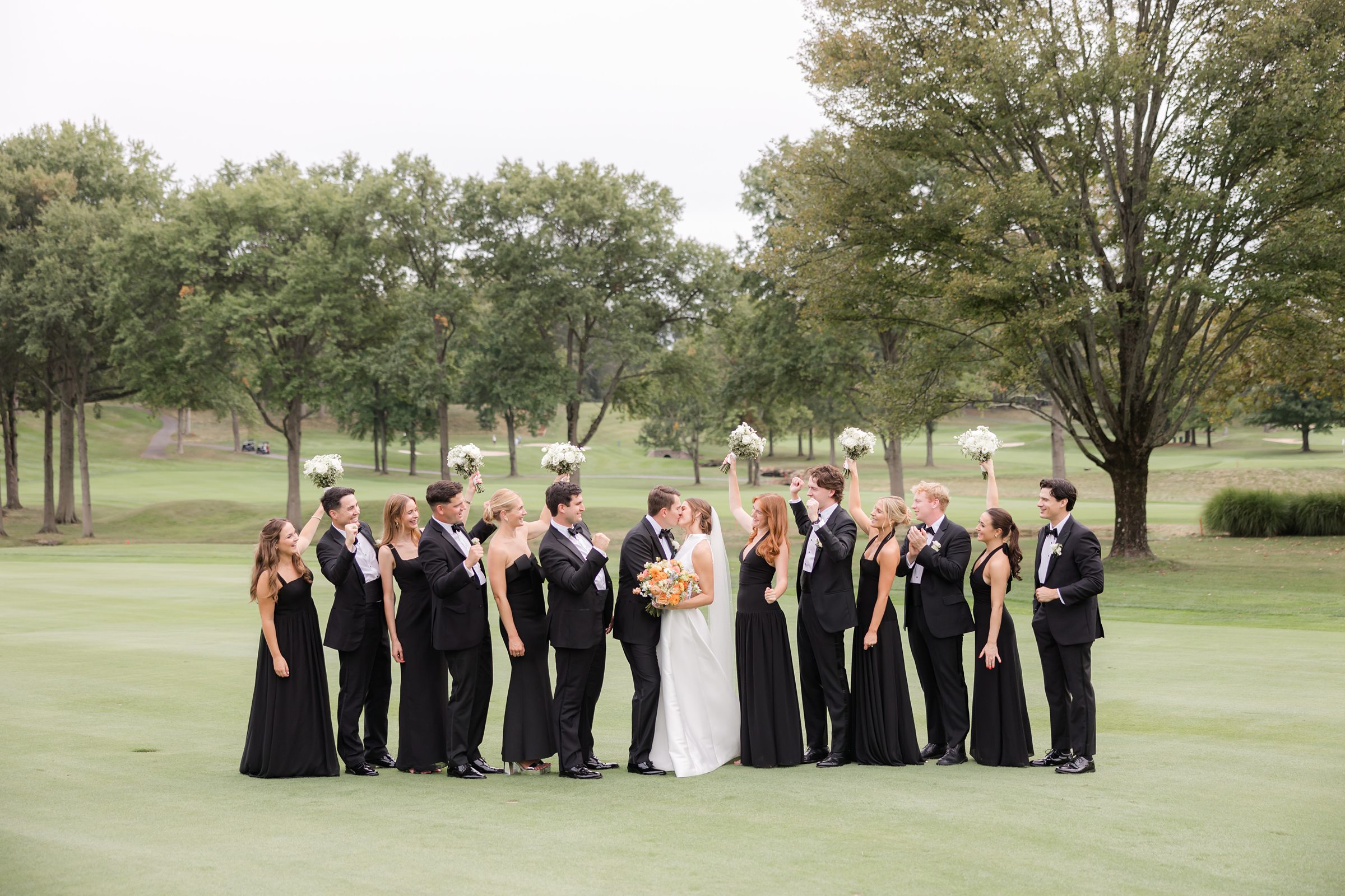 Bride and groom stand surrounded by their bridesmaids and groomsmen on a lush green lawn, sharing smiles and laughter together.