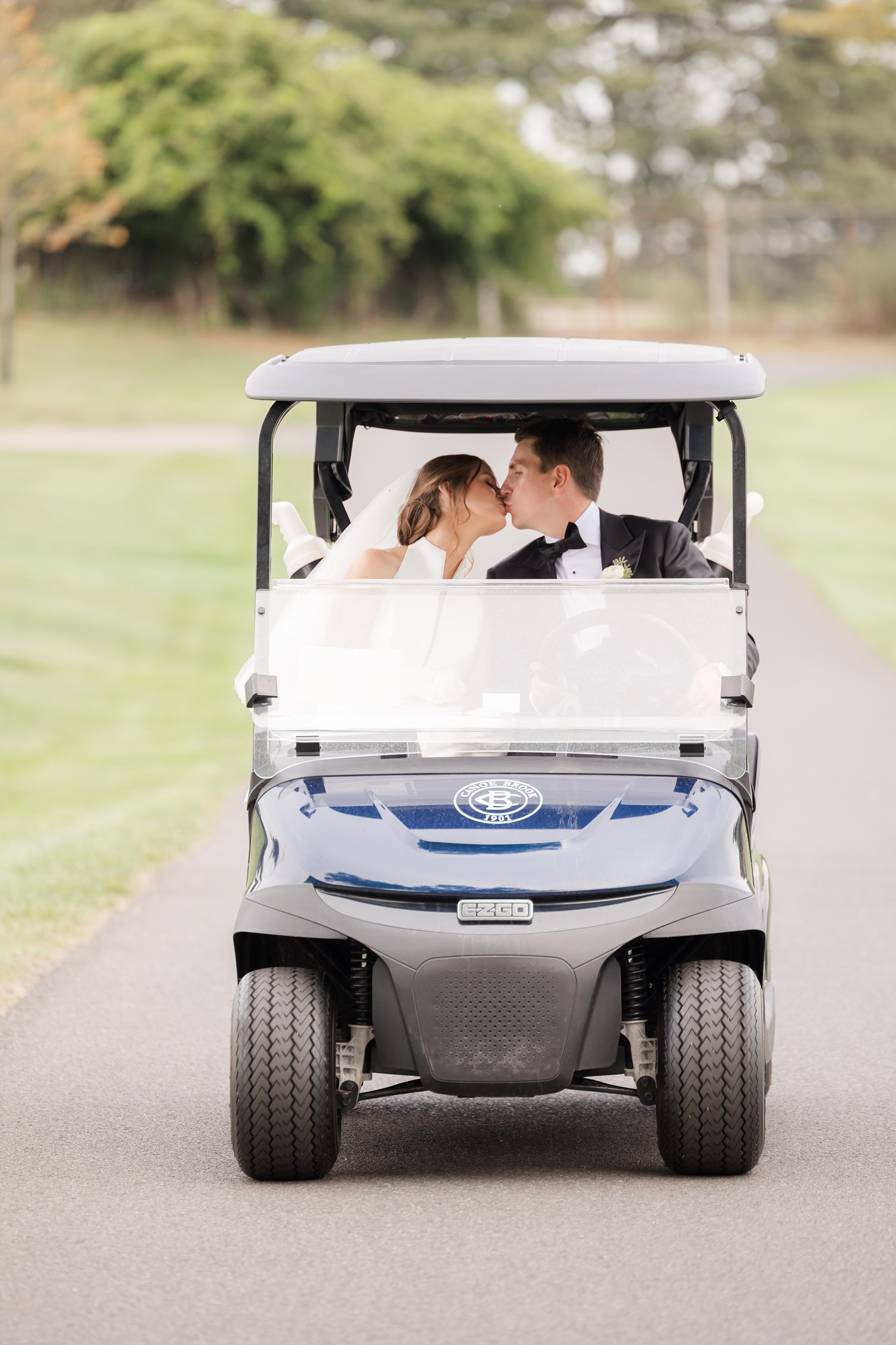Bride and groom share a quiet kiss in a golf cart, surrounded by soft greenery.