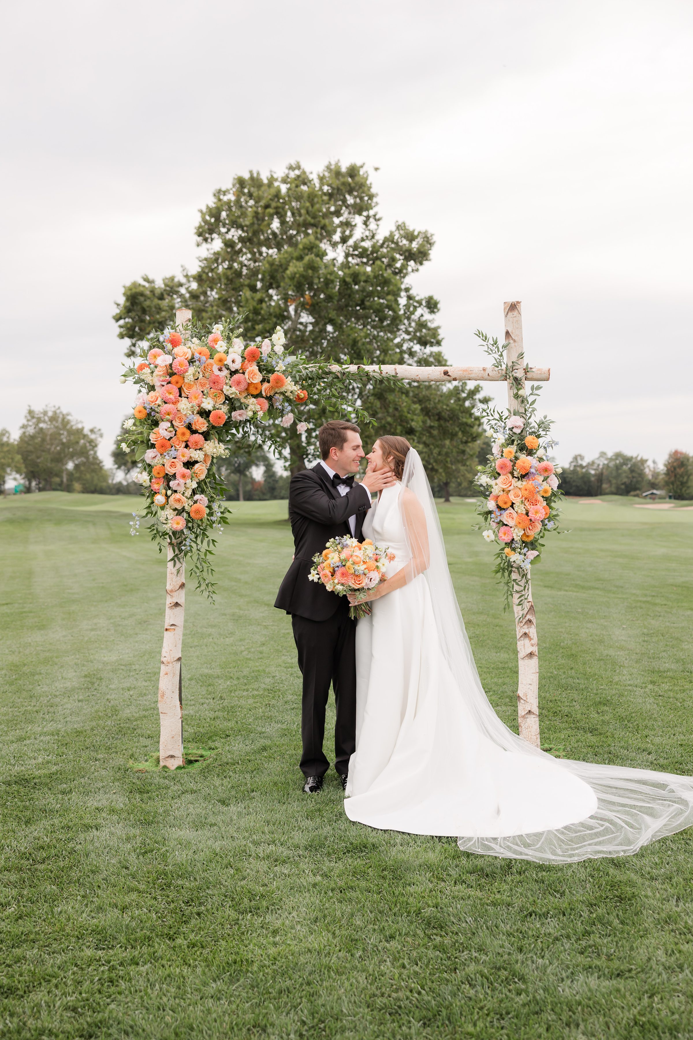 Beneath a floral arch in an open field, the groom gently cups his bride’s face as they share a quiet, loving moment together.