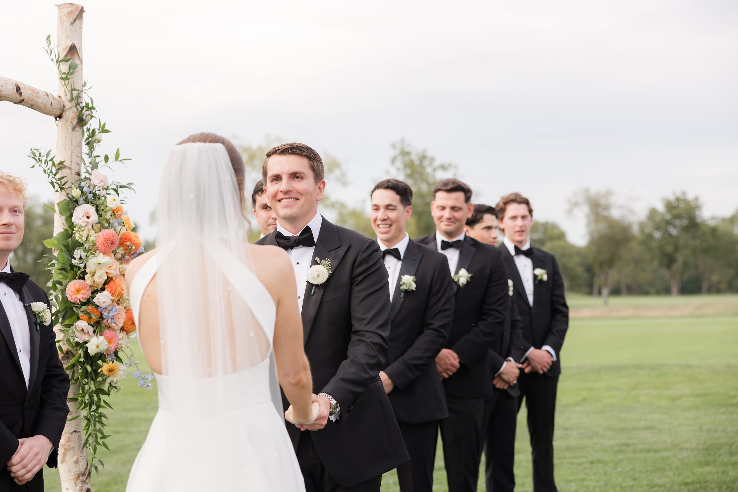 Groom beams as he takes the bride’s hands at the altar, surrounded by his groomsmen on a green lawn.