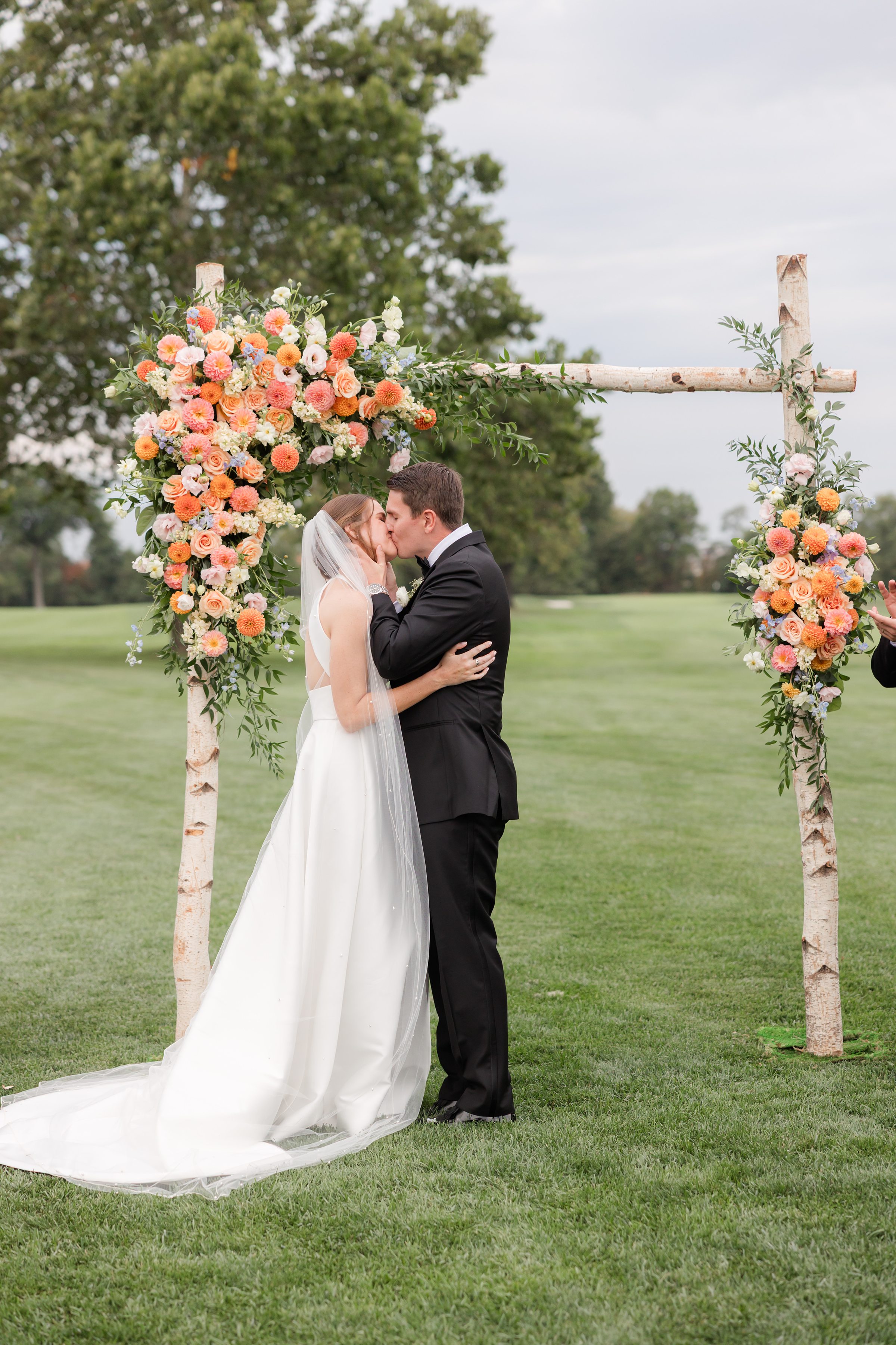 Bride and groom share a kiss beneath a floral ceremony arch on a green lawn during their outdoor wedding.