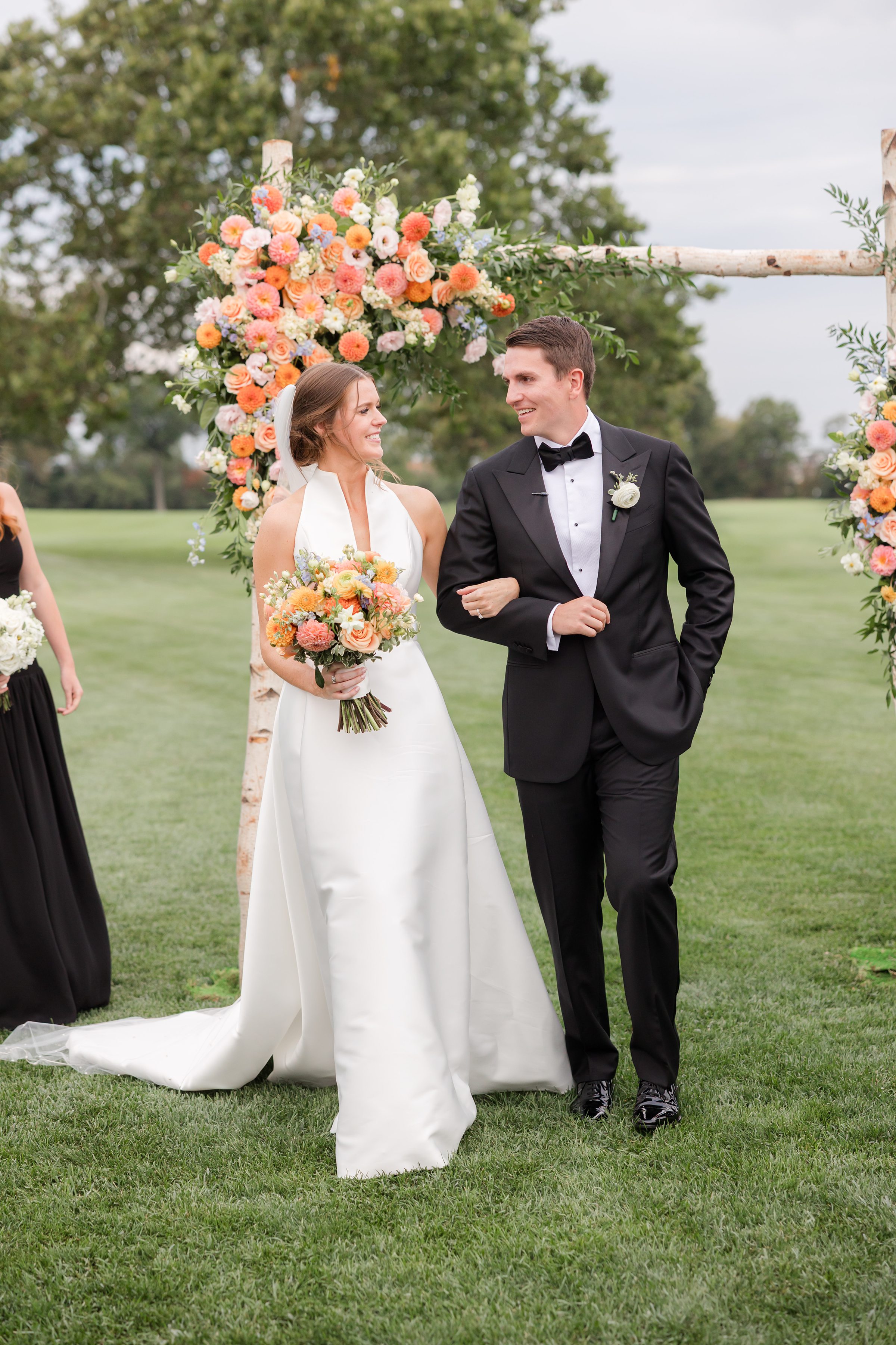 Bride and groom walk together beneath a floral arch, smiling at each other on a green lawn.