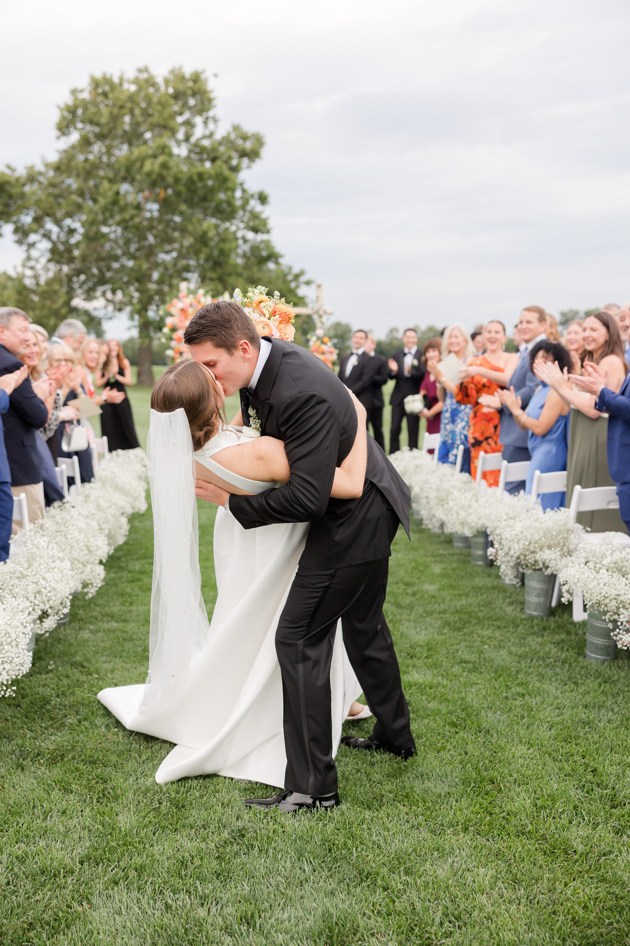 Groom dips the bride into a kiss in the aisle as guests stand and applaud around them during their outdoor wedding ceremony.