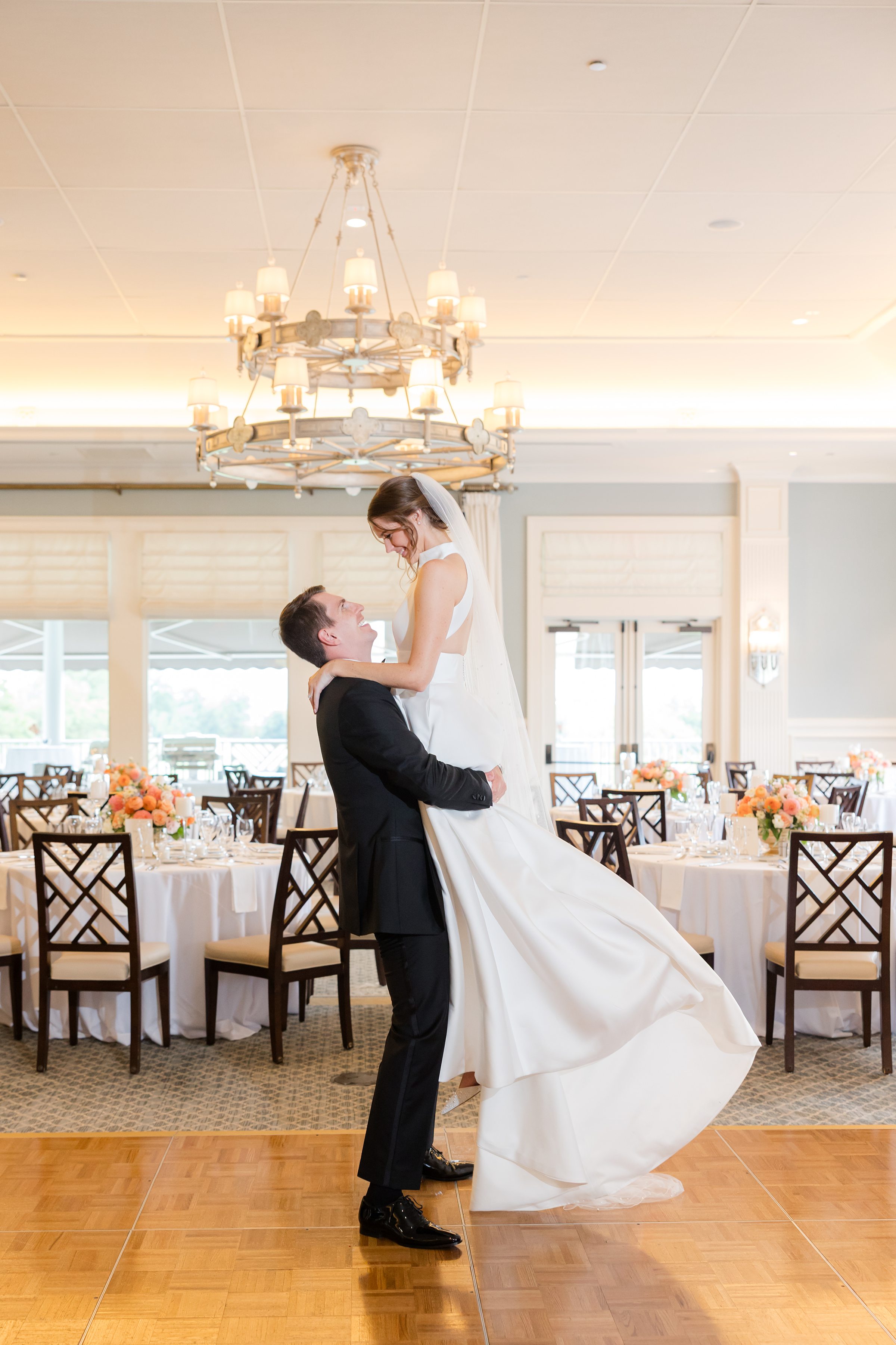 Groom lifts the bride into the air on the dance floor, smiling up at her in a bright, elegant reception space.