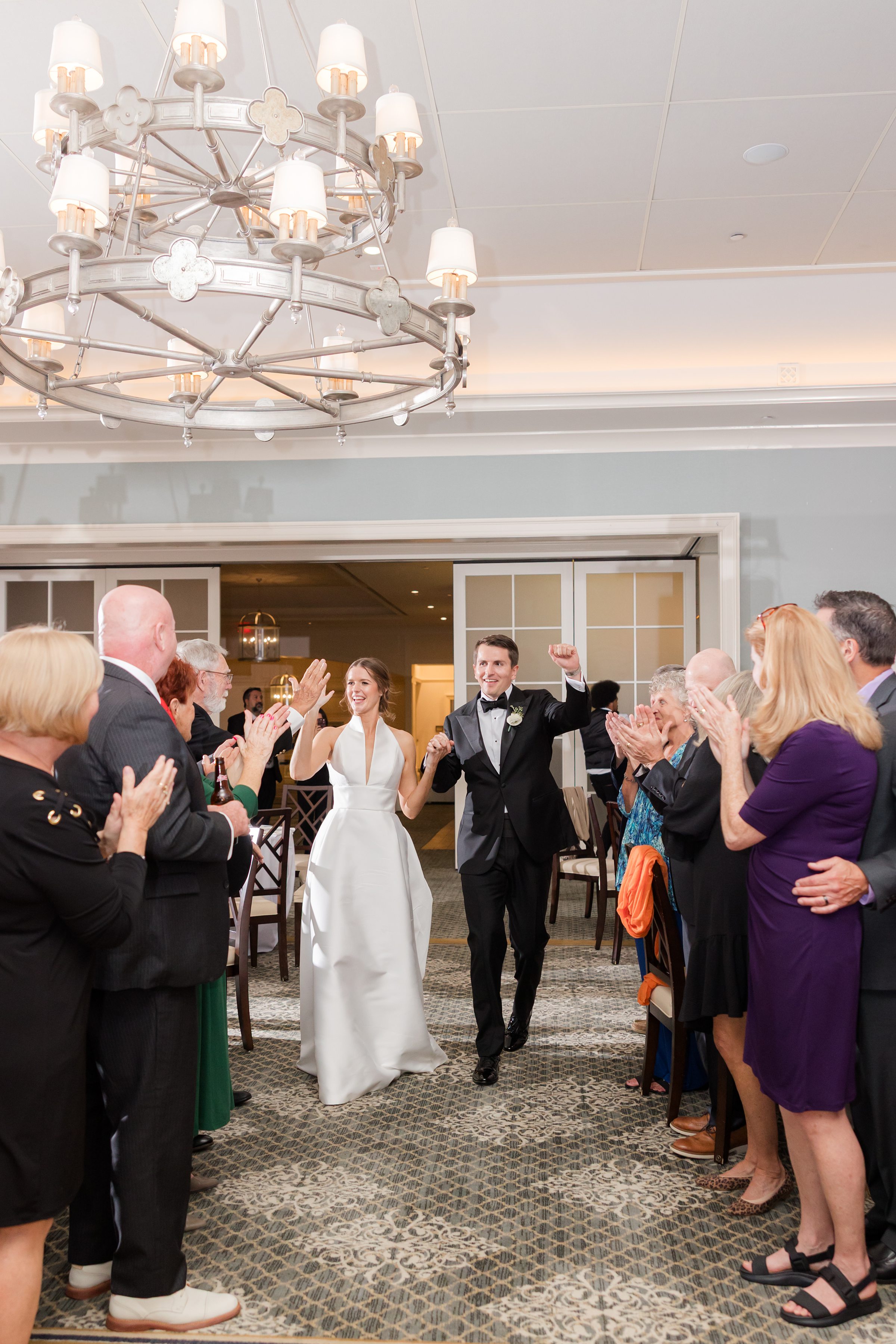 Bride and groom make their grand entrance as guests cheer and clap beneath a glowing chandelier.