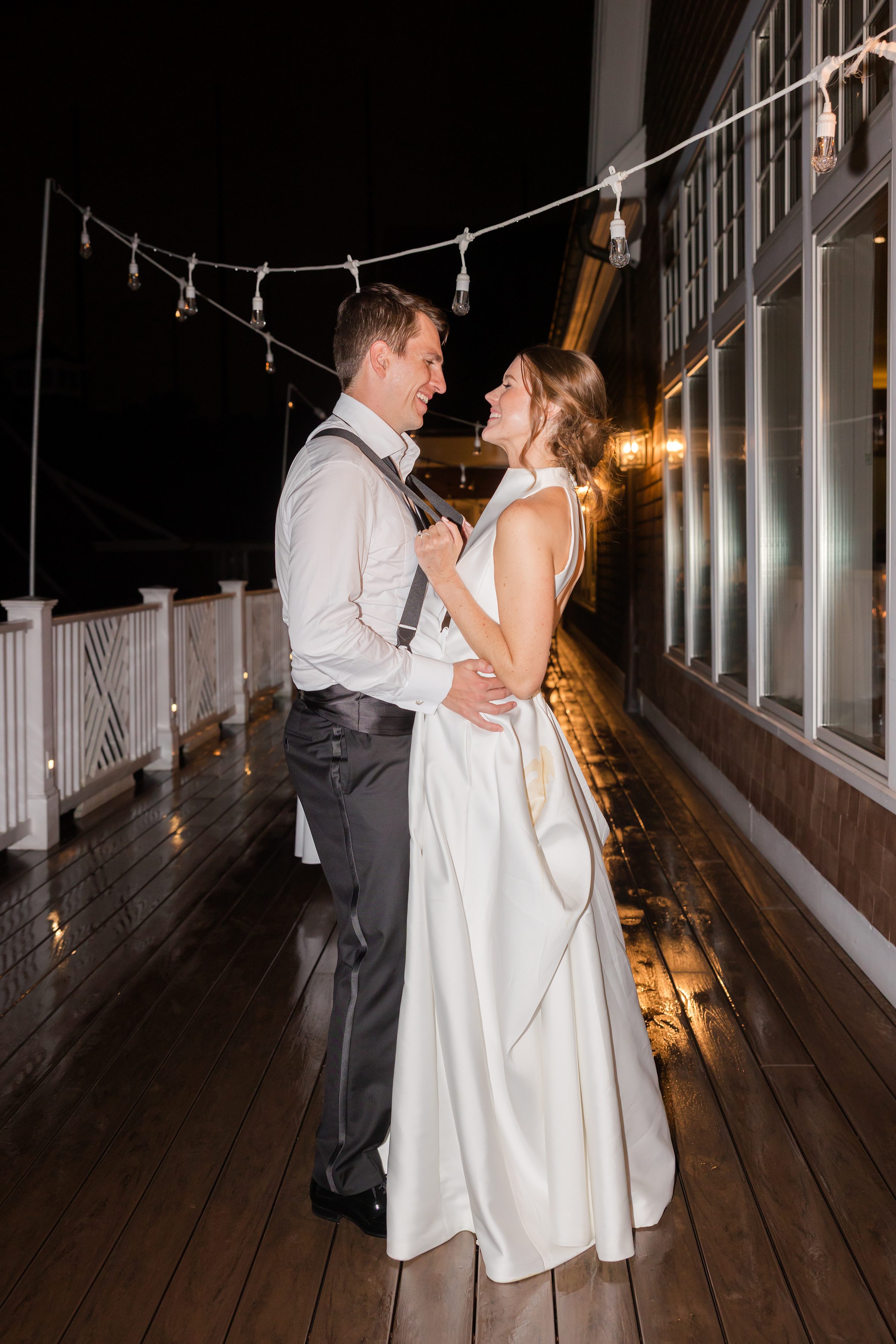 Bride and groom stand close together on a softly lit terrace at night, smiling at each other beneath string lights.
