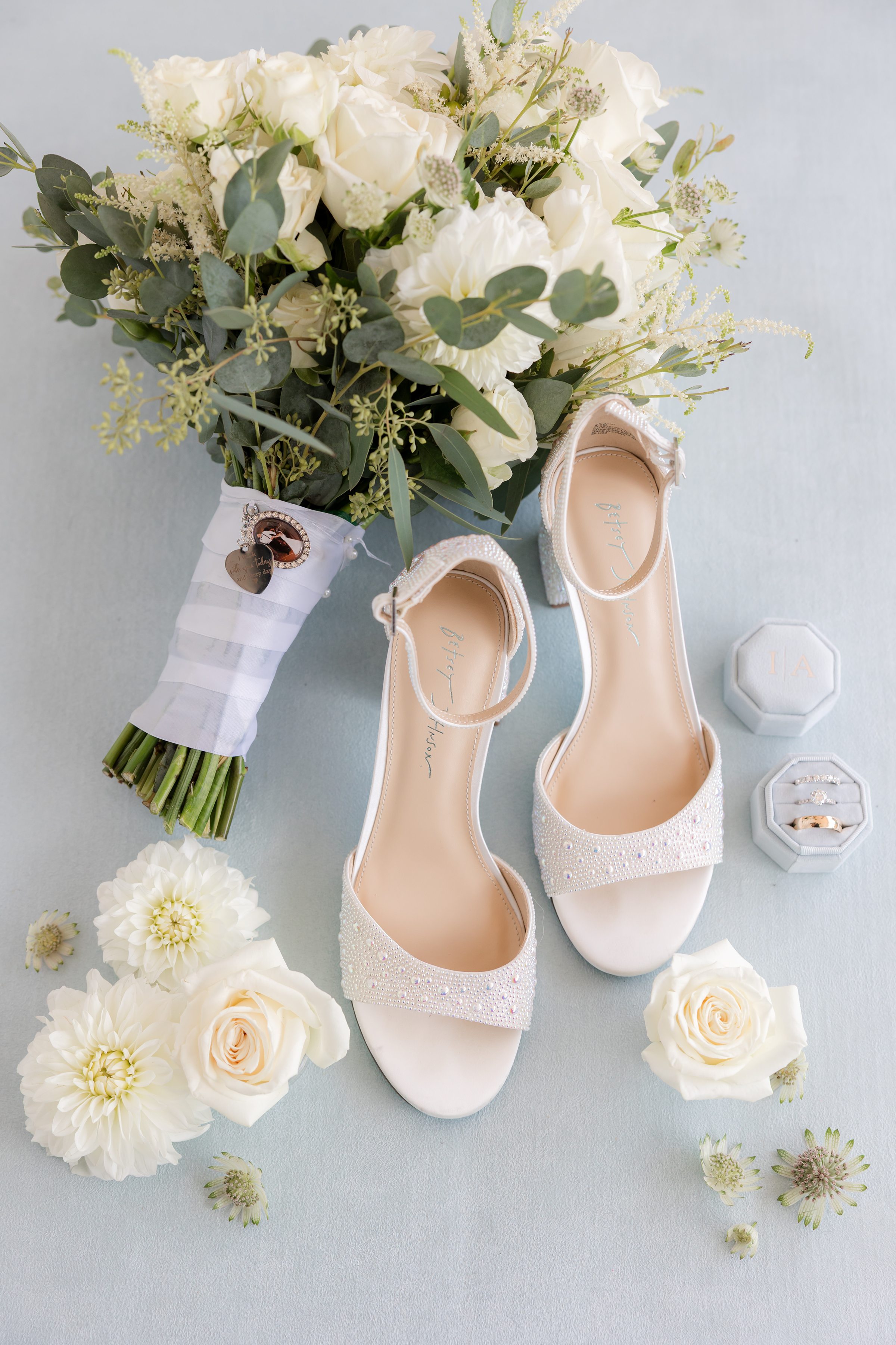 White bridal sandals, bouquet of white flowers, and ring boxes arranged on a light blue surface.