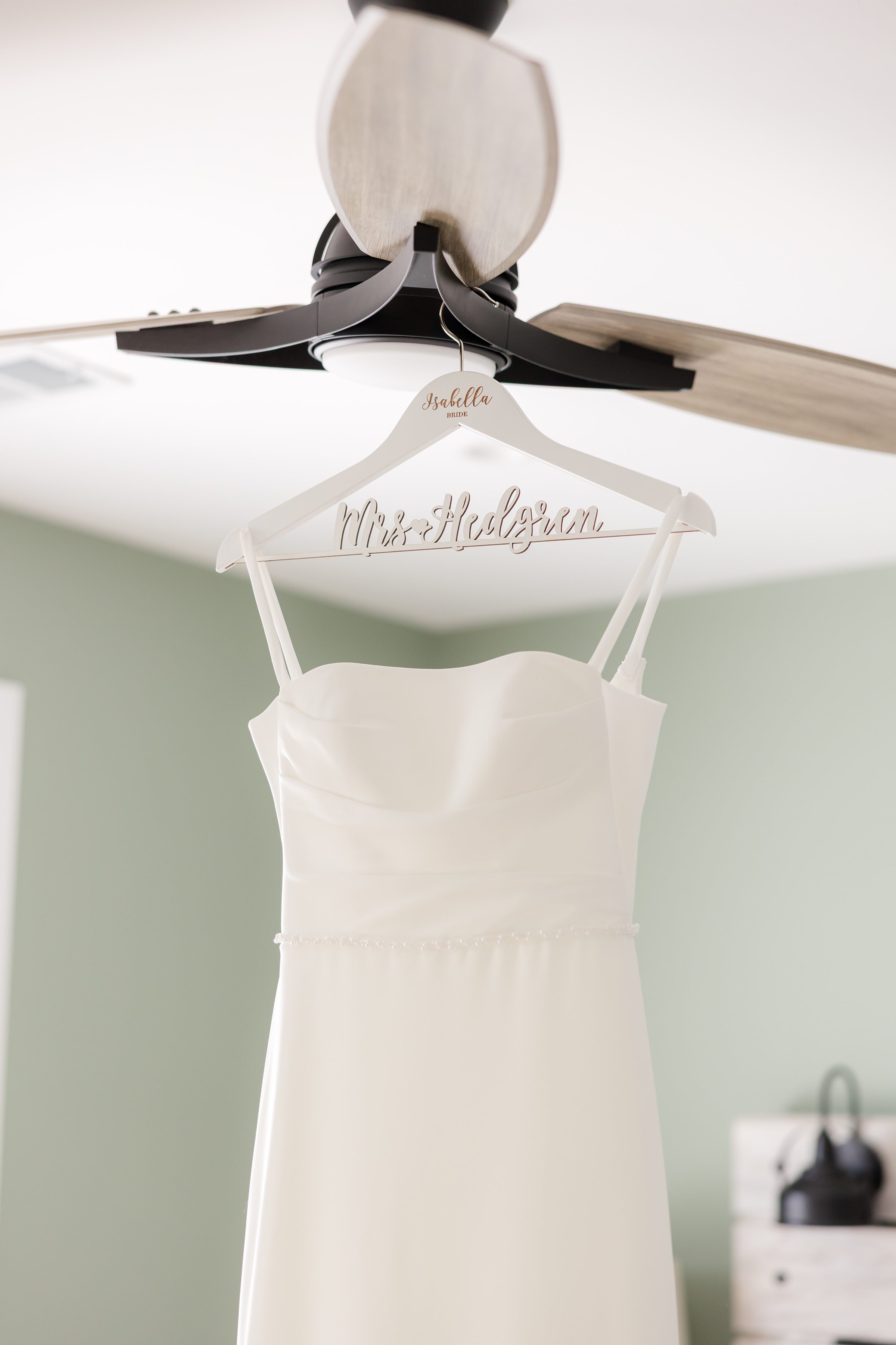 Simple white wedding dress hanging from a personalized hanger beneath a ceiling fan in a softly lit room.