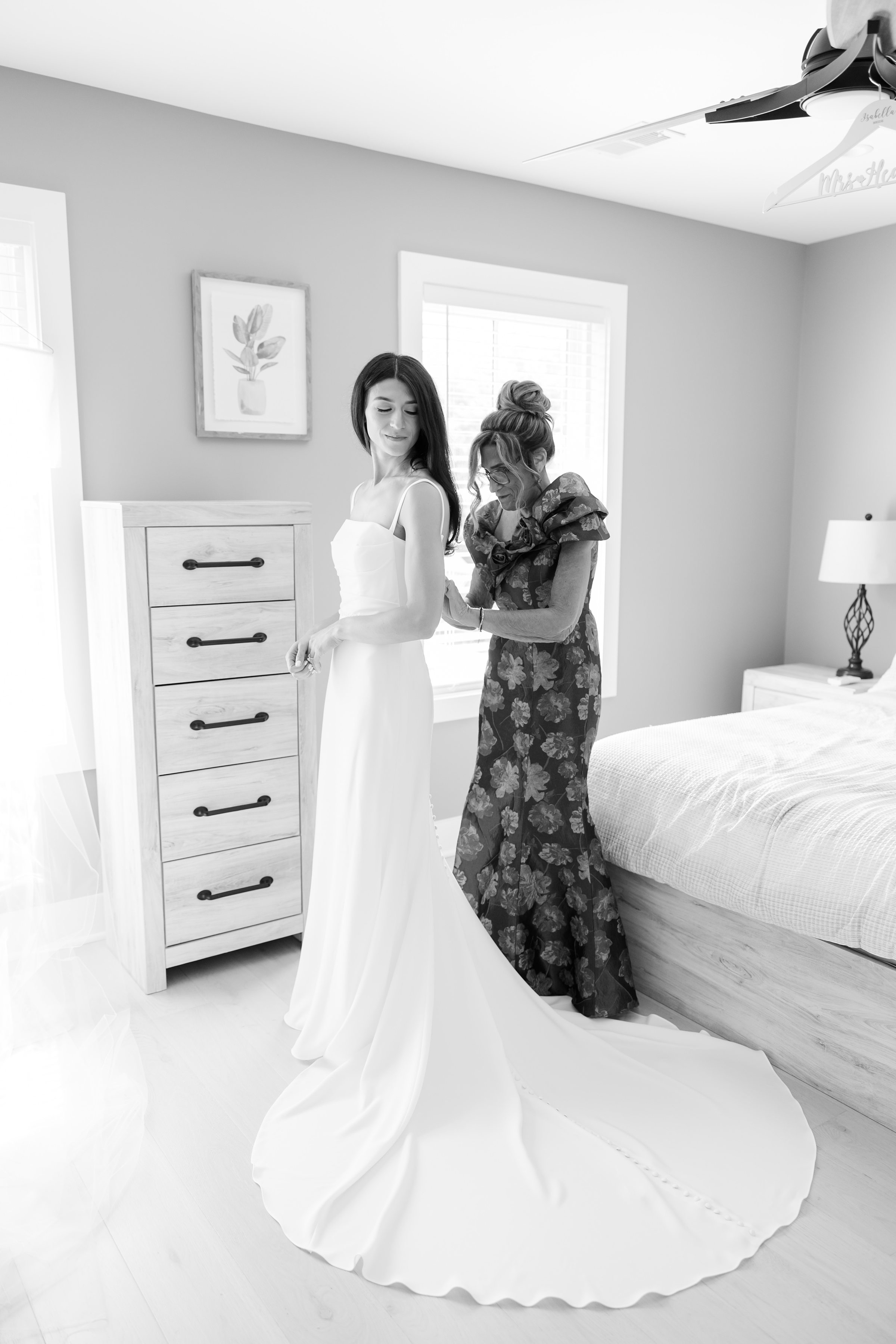 Mother gently fastens her daughter’s wedding dress as the bride looks over her shoulder with a soft, emotional smile, sharing a quiet, intimate moment before the ceremony.